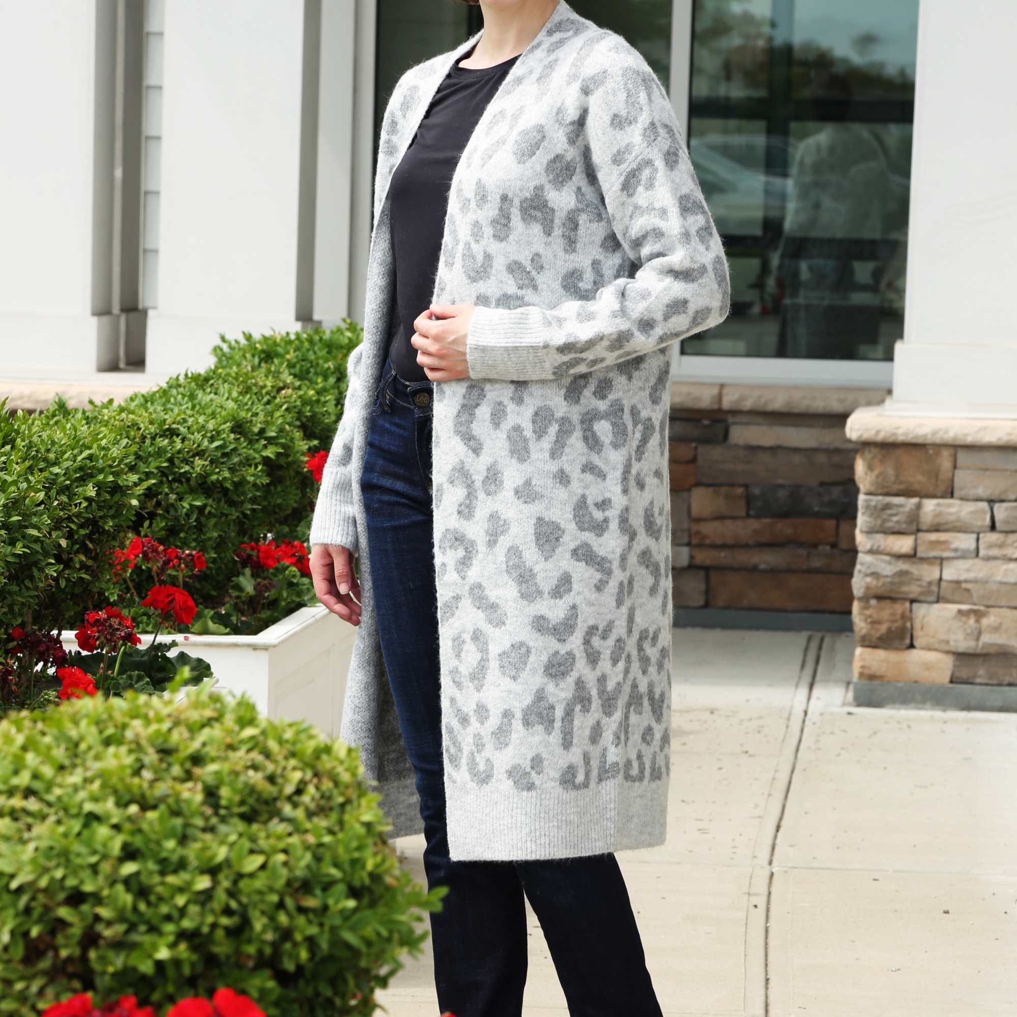 Person in a long gray leopard-print cardigan and dark jeans standing by red flowers outside a building.