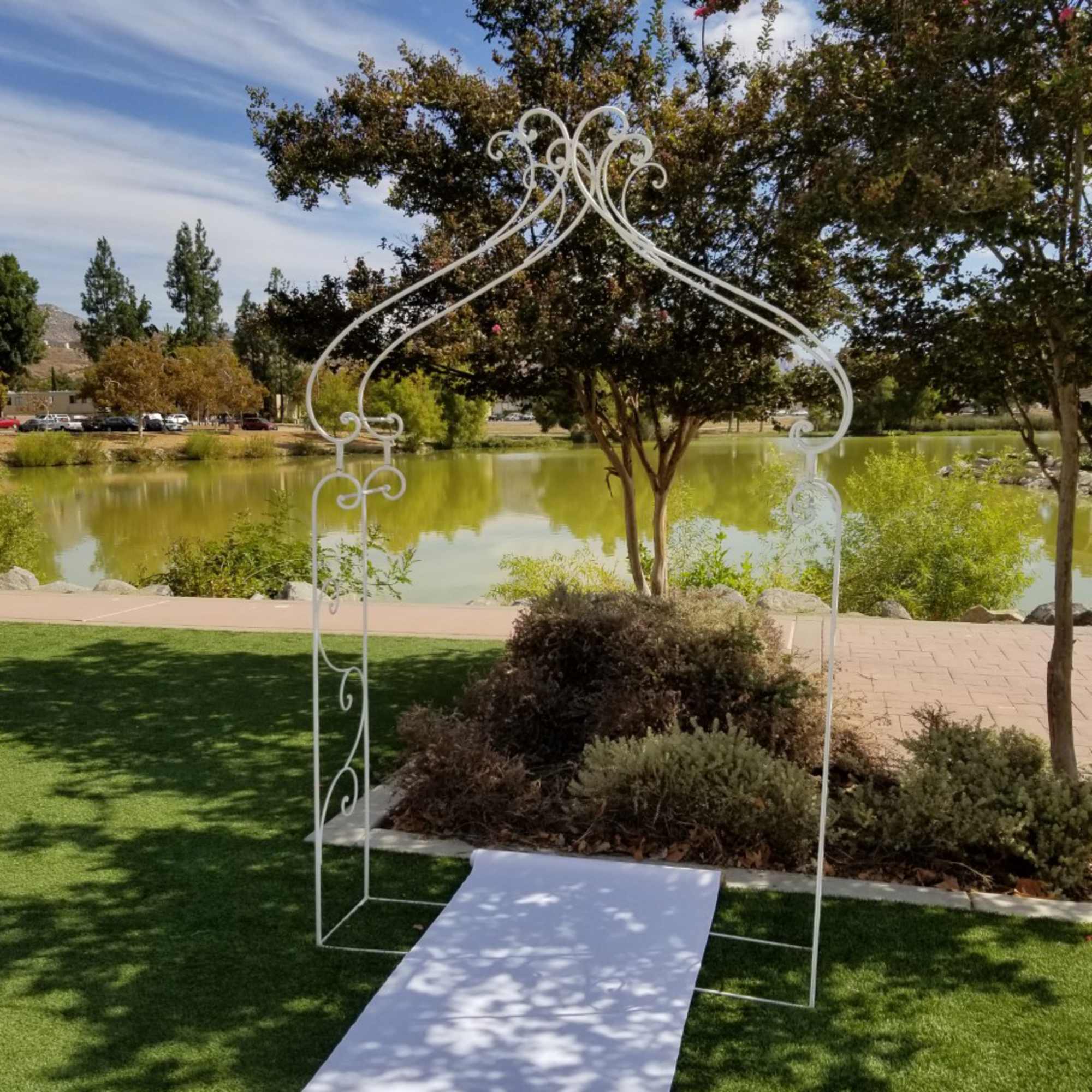 White decorative wedding arch over a white aisle runner by a pond, photo 2 of 3