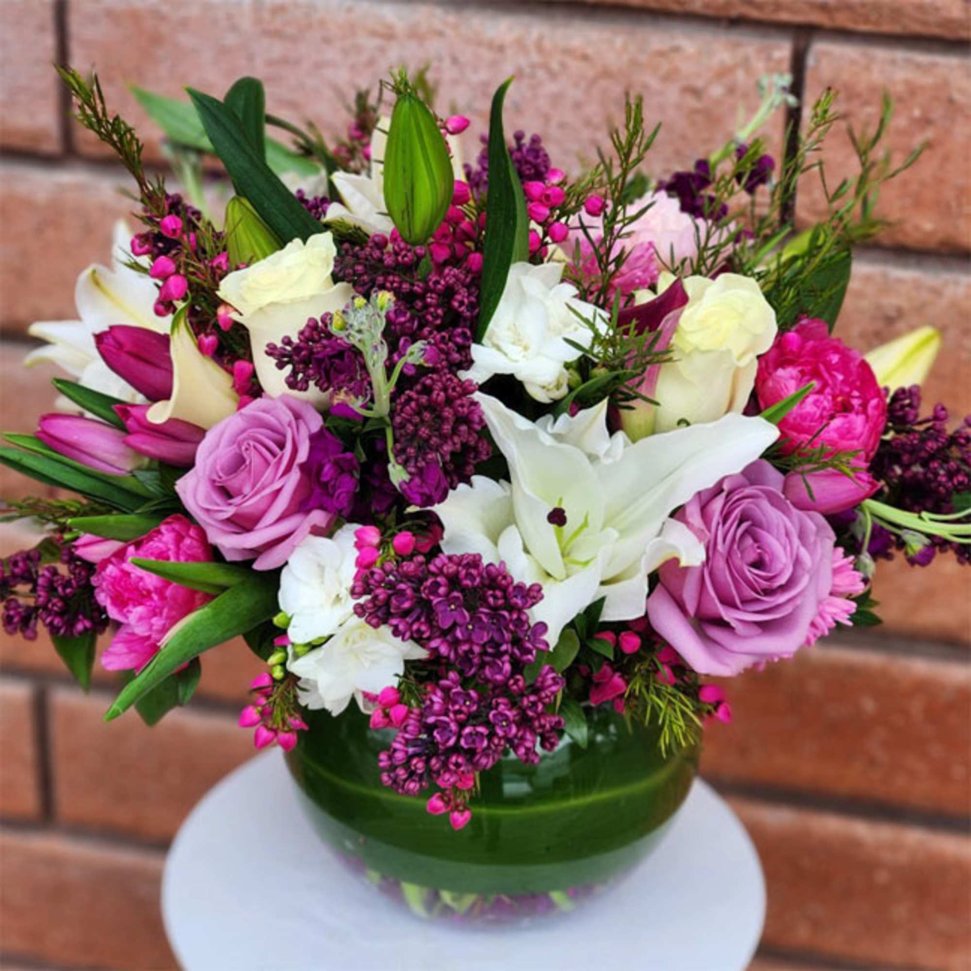Round arrangement of purple, pink, and white flowers in a green-wrapped glass bowl vase
