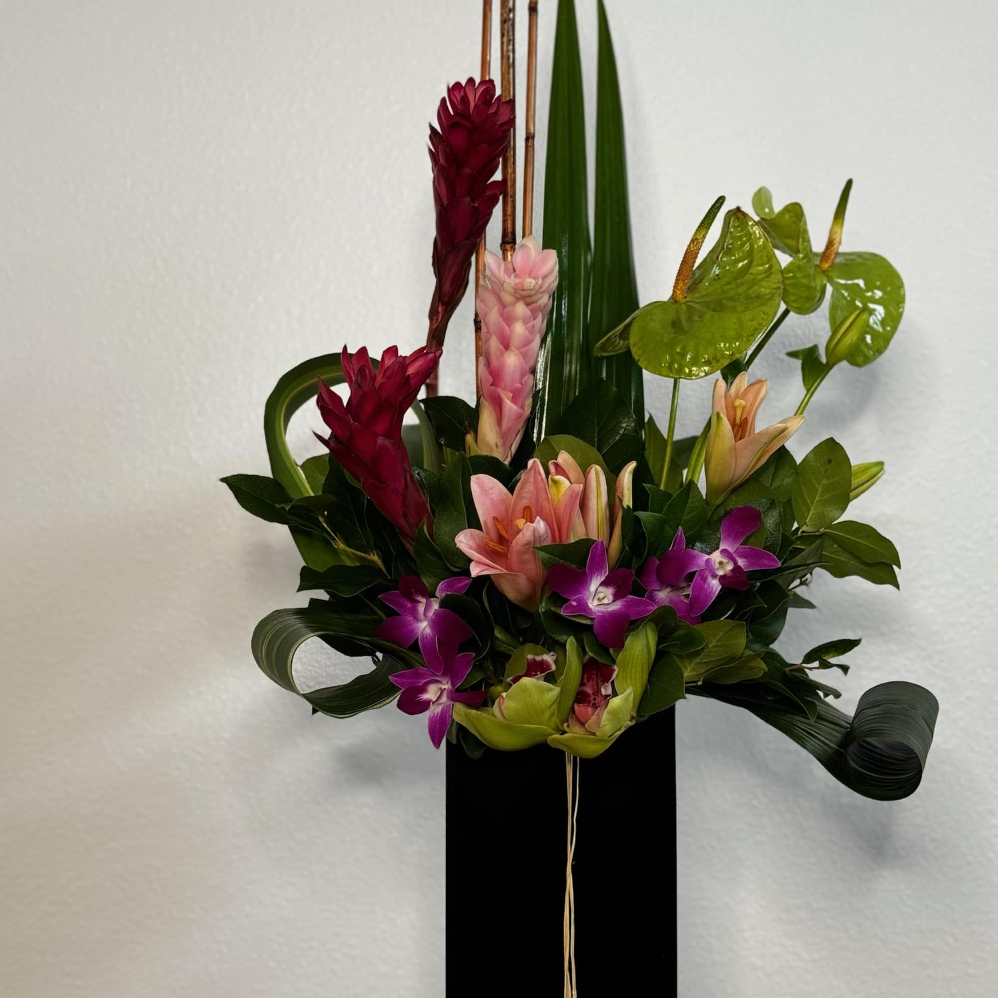 Tall tropical arrangement with pink lilies, purple orchids, and green anthuriums in a black rectangular vase