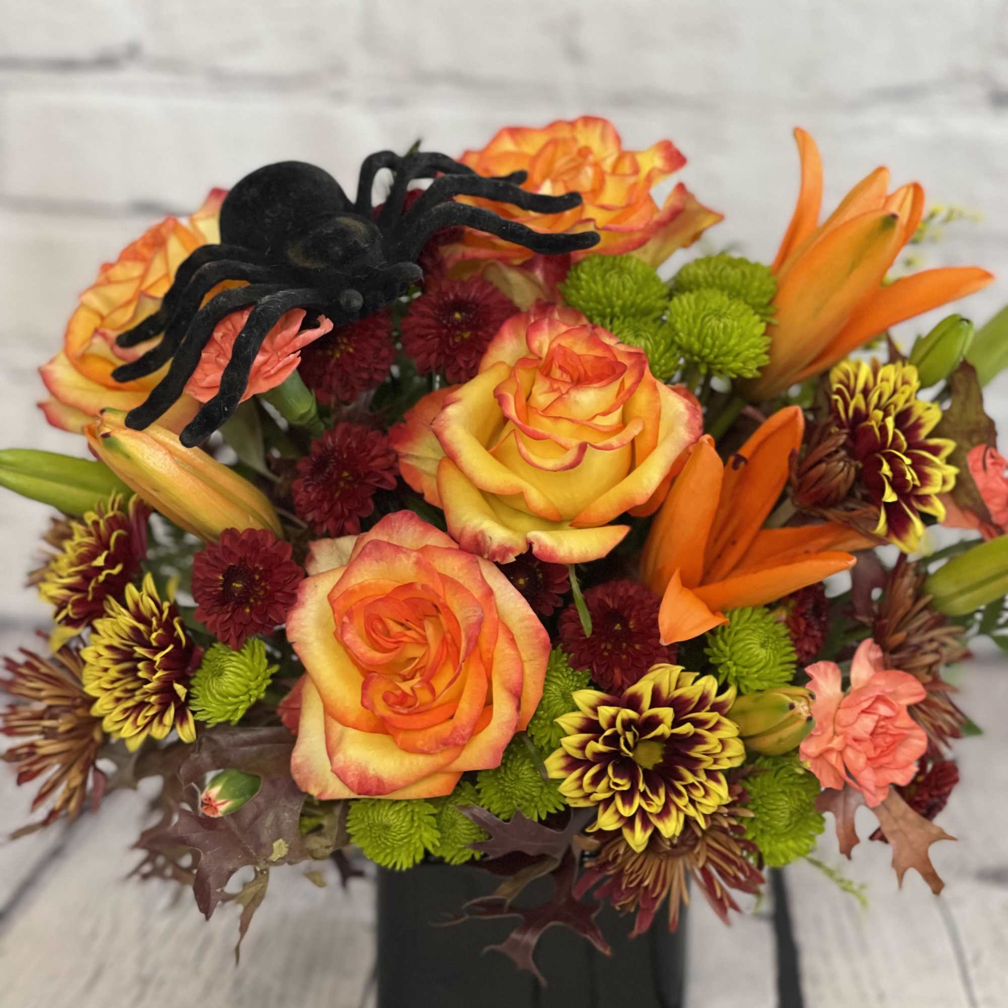 Orange roses and mixed mums in a black vase with a decorative spider accent