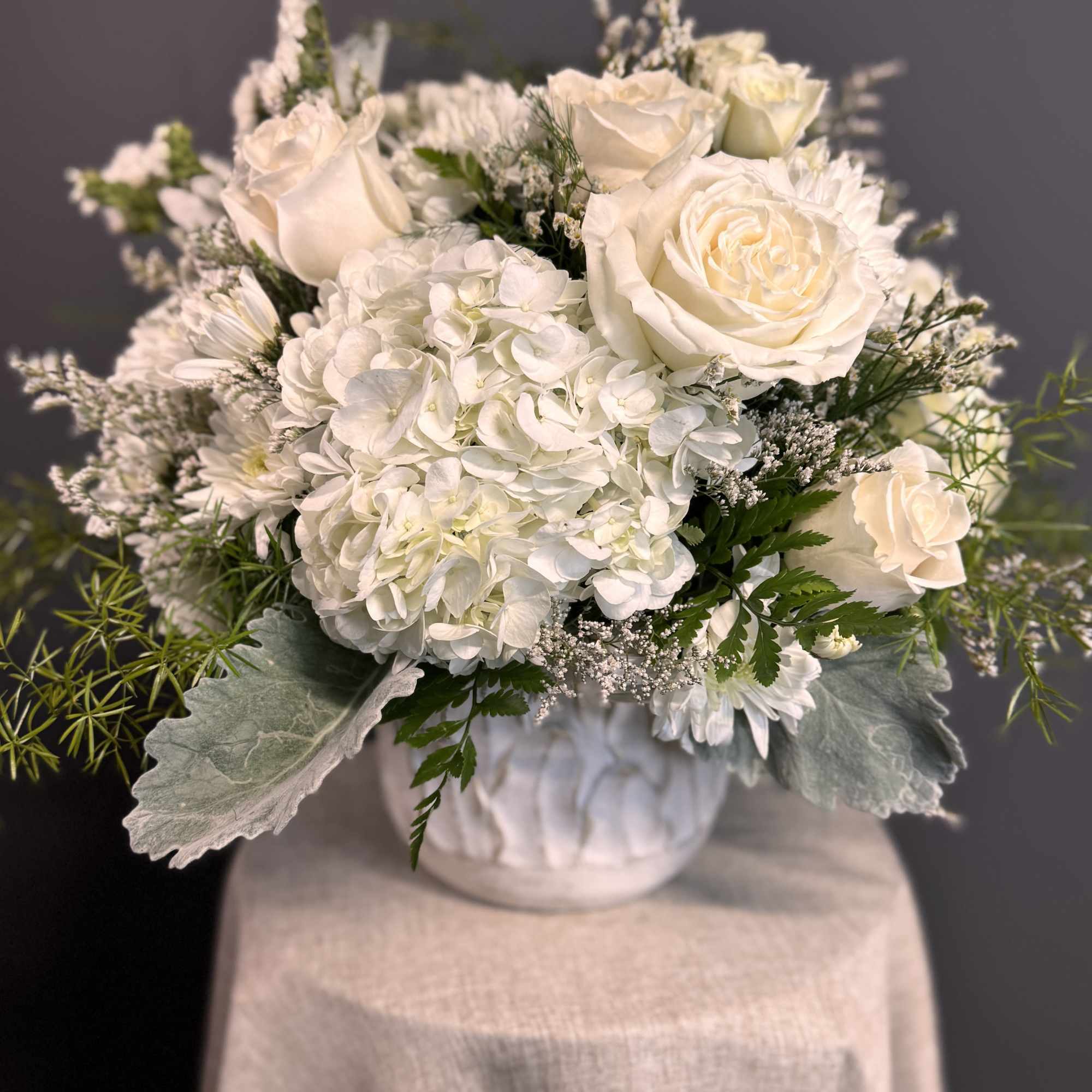 All-white arrangement of roses, hydrangeas, and mums in a white ceramic vase on a draped pedestal.