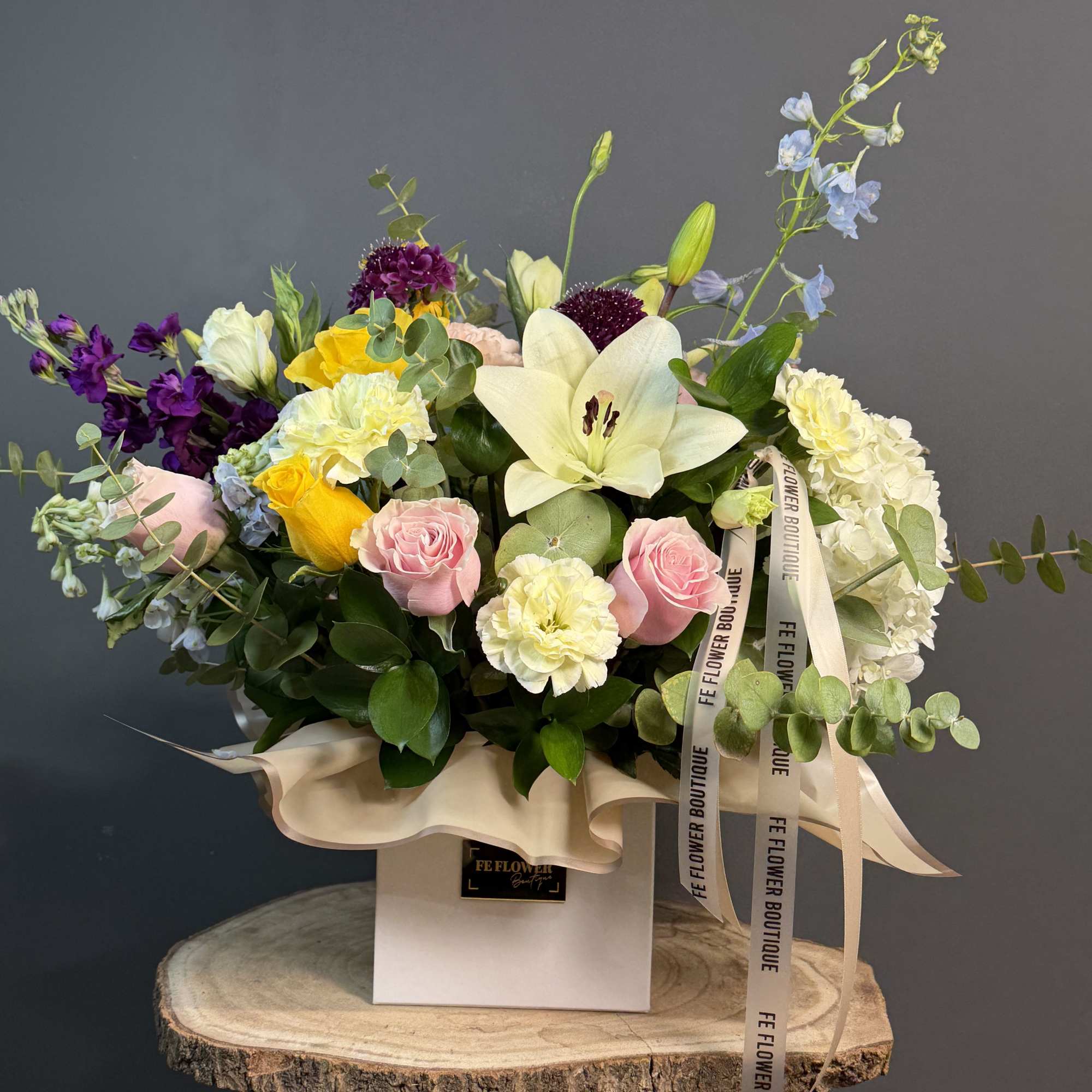 Colorful mixed flower arrangement with lilies, roses, and carnations in a white box on a wood stand.