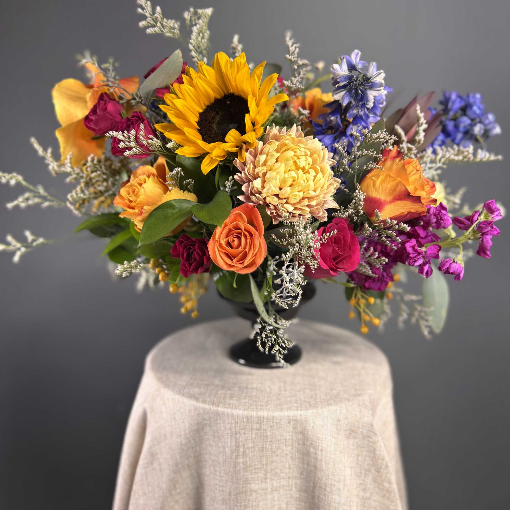 Bright mixed arrangement with sunflower, orange roses, and colorful blooms in a black pedestal vase