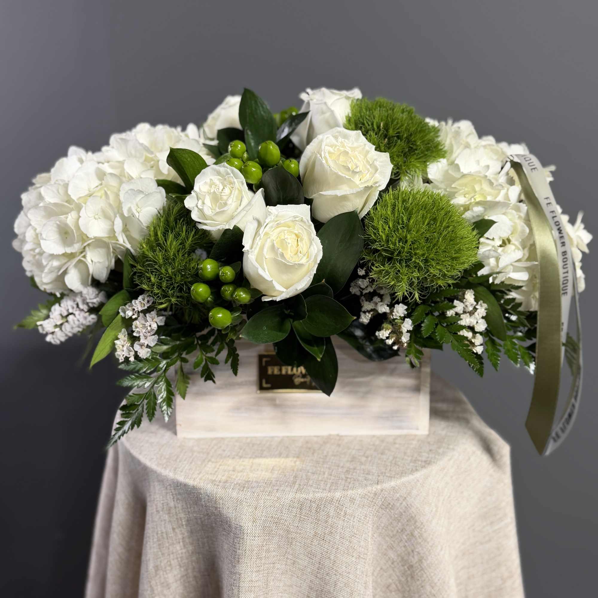 Low white arrangement of roses, hydrangeas, and green dianthus in a rectangular wooden box.