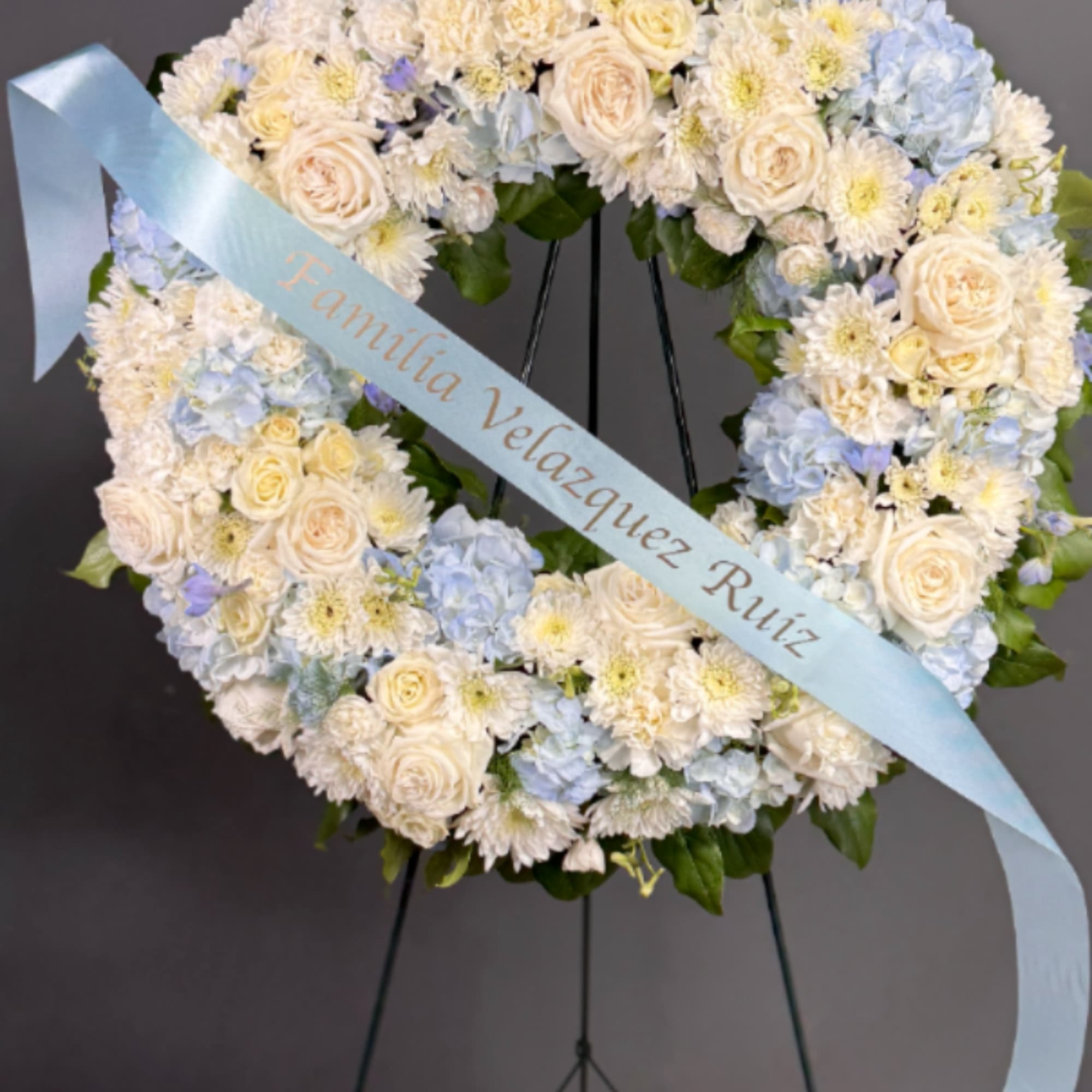 Standing wreath of white and pale blue flowers with a light blue message ribbon on an easel.