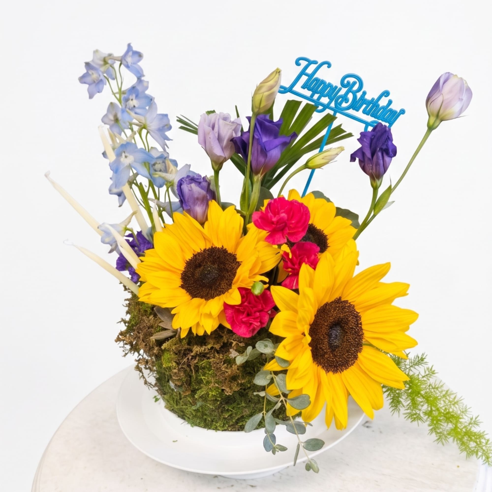 Sunflower arrangement with purple and blue blooms in a moss-covered container