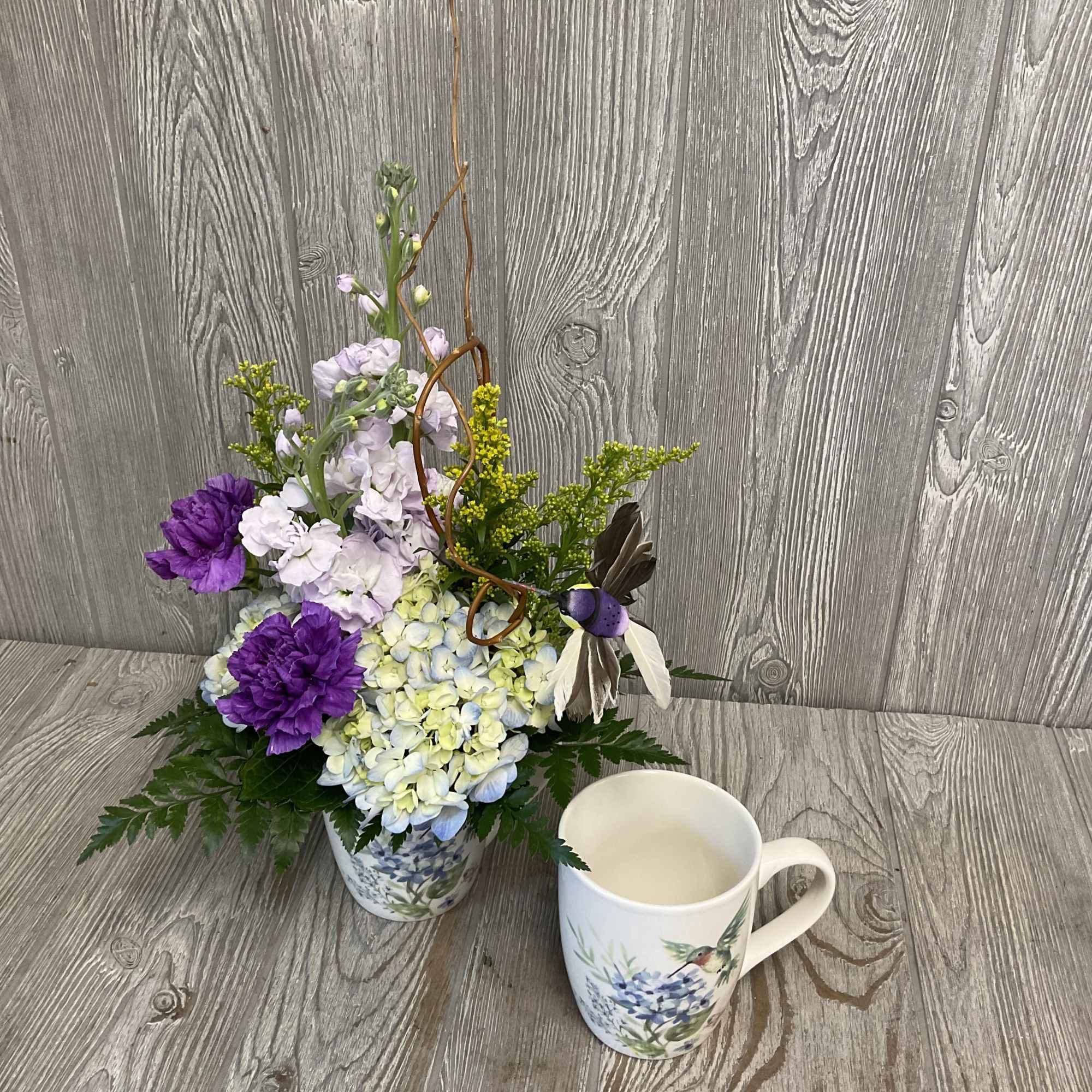 Purple and white floral arrangement in a patterned ceramic pot beside a matching mug