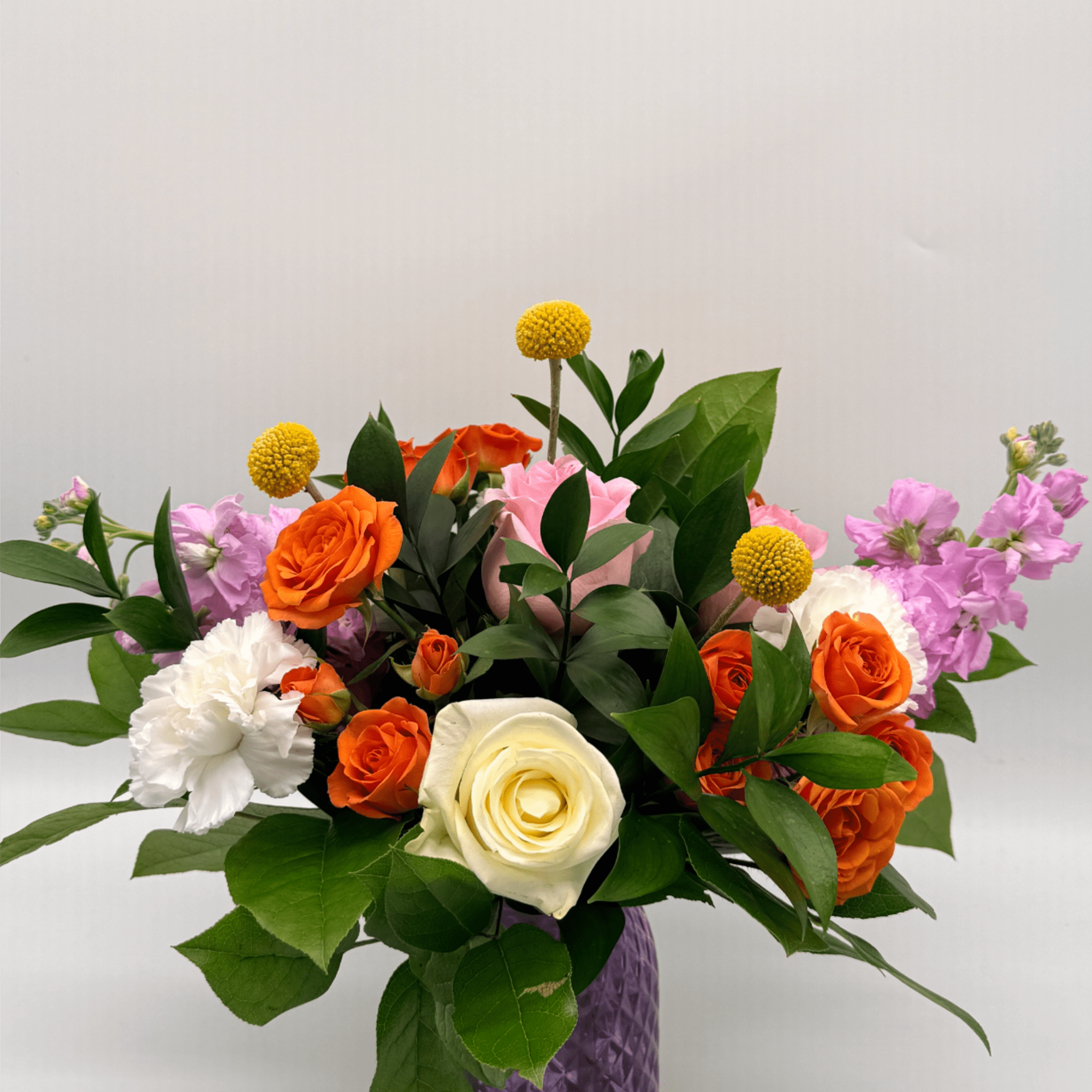 Colorful arrangement of orange roses, white rose and carnation, and pink blooms in a purple glass vase