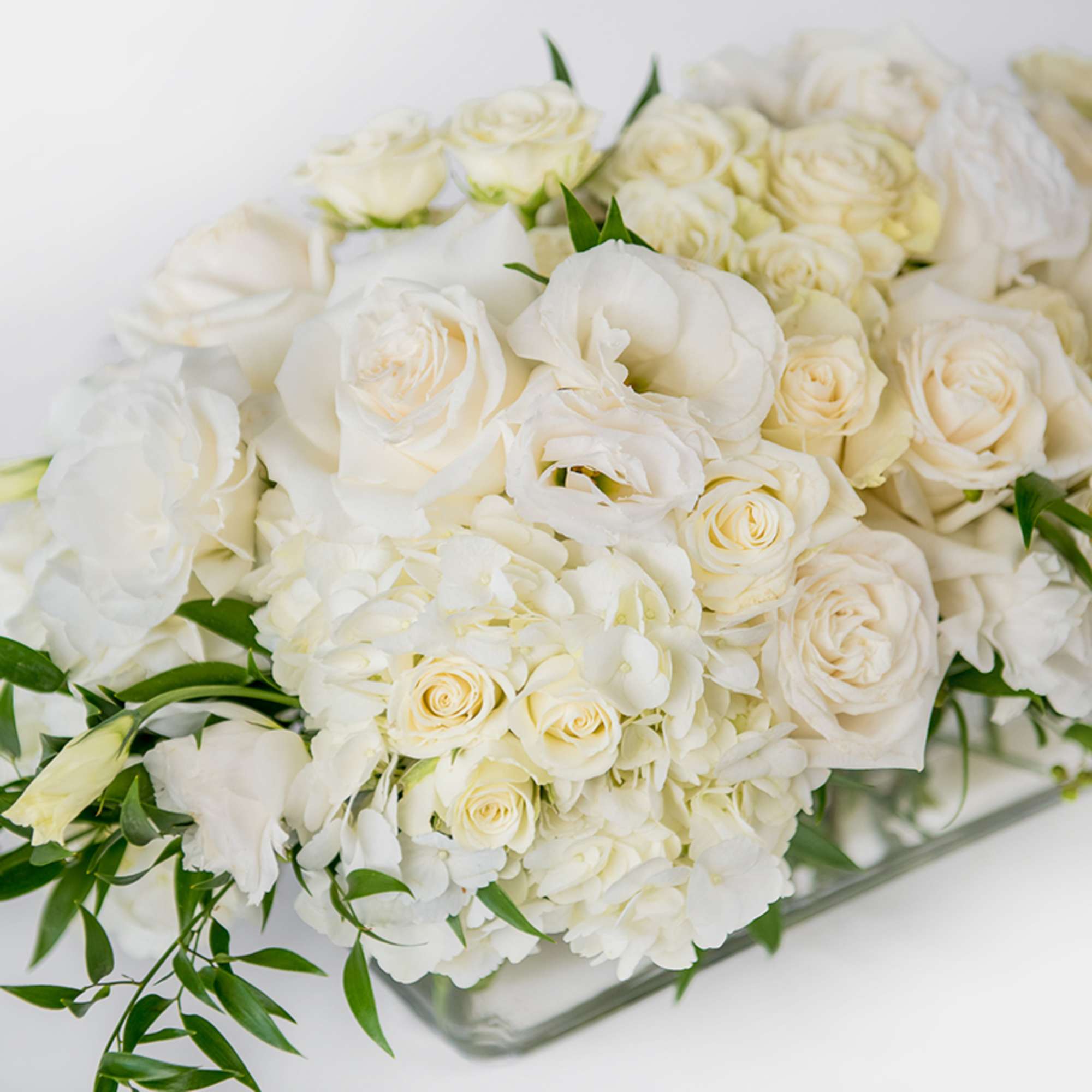Low arrangement of white and cream roses, lisianthus, and hydrangeas in a clear glass rectangular vase