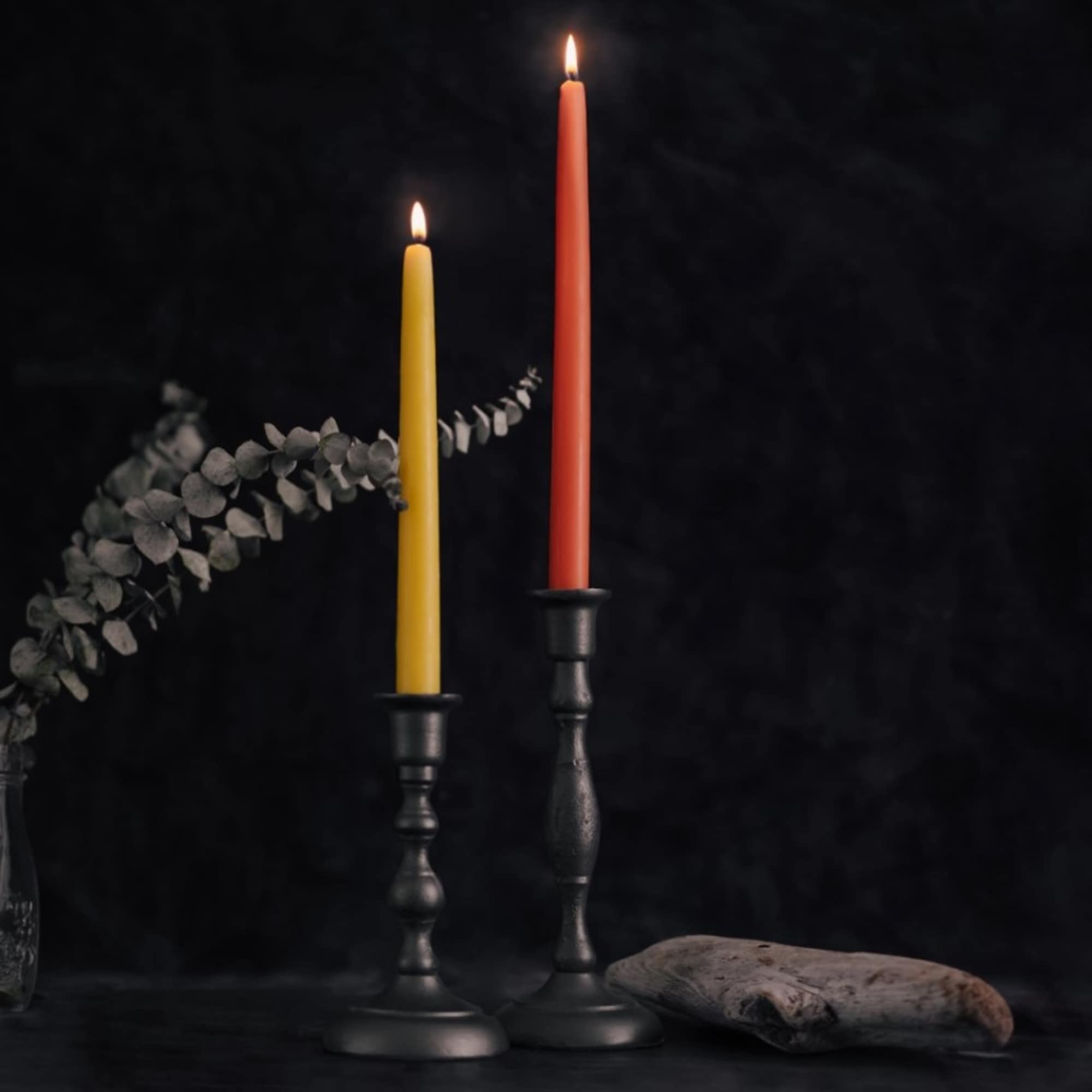 Two lit yellow and orange taper candles in dark metal holders against a black background