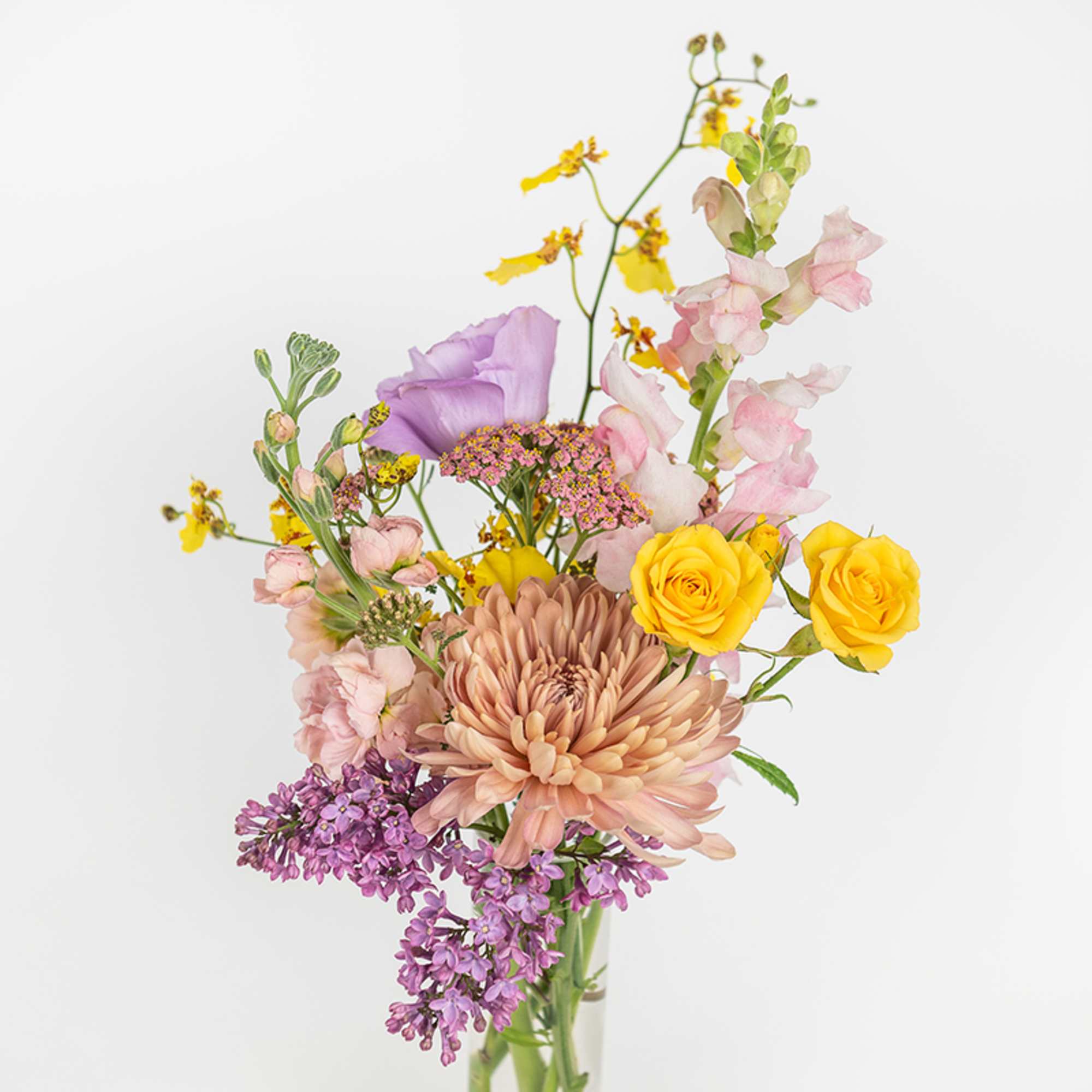 Loose bouquet with yellow roses, peach chrysanthemum, lilac, and pastel blooms in a clear glass vase