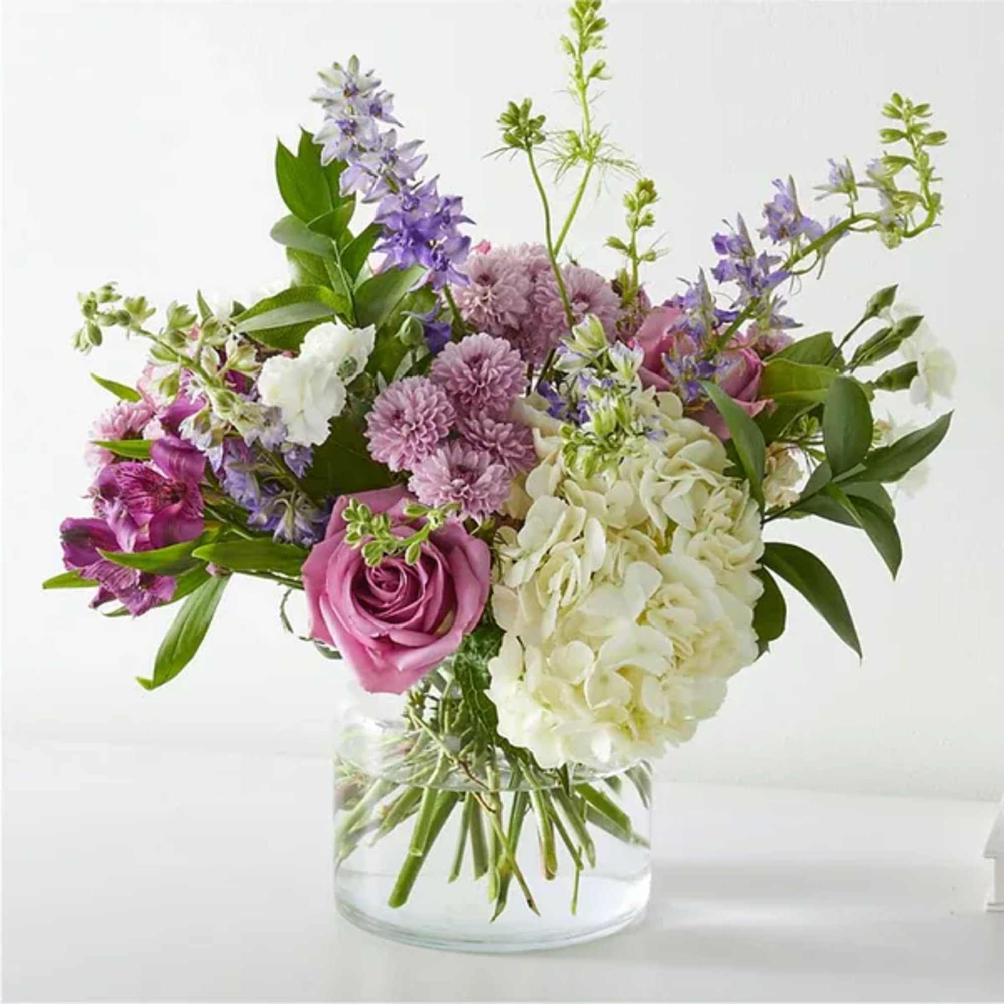 Arrangement of white hydrangeas, lavender roses, and purple blooms in a clear glass vase