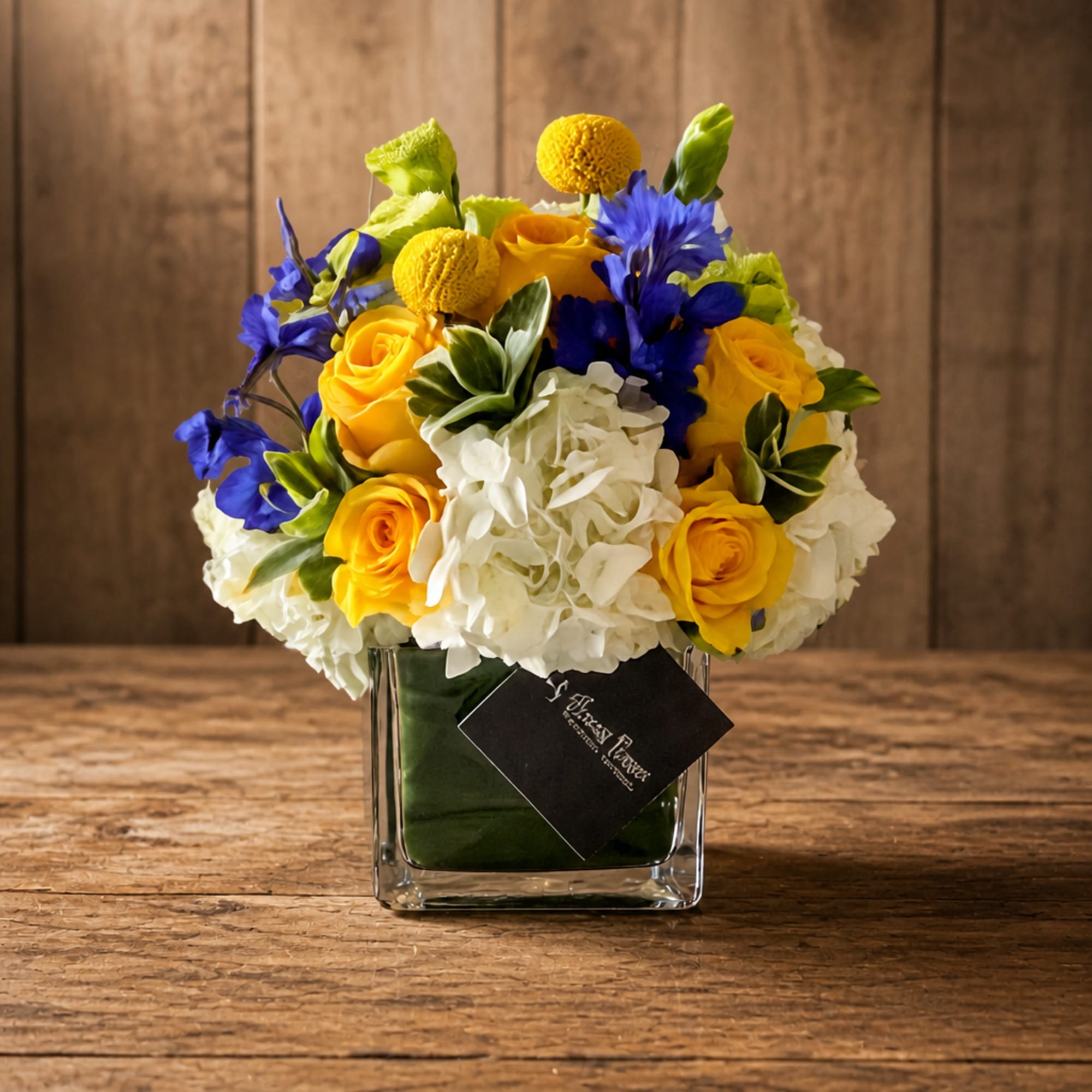 Yellow roses, white hydrangeas, and blue flowers in a square glass vase