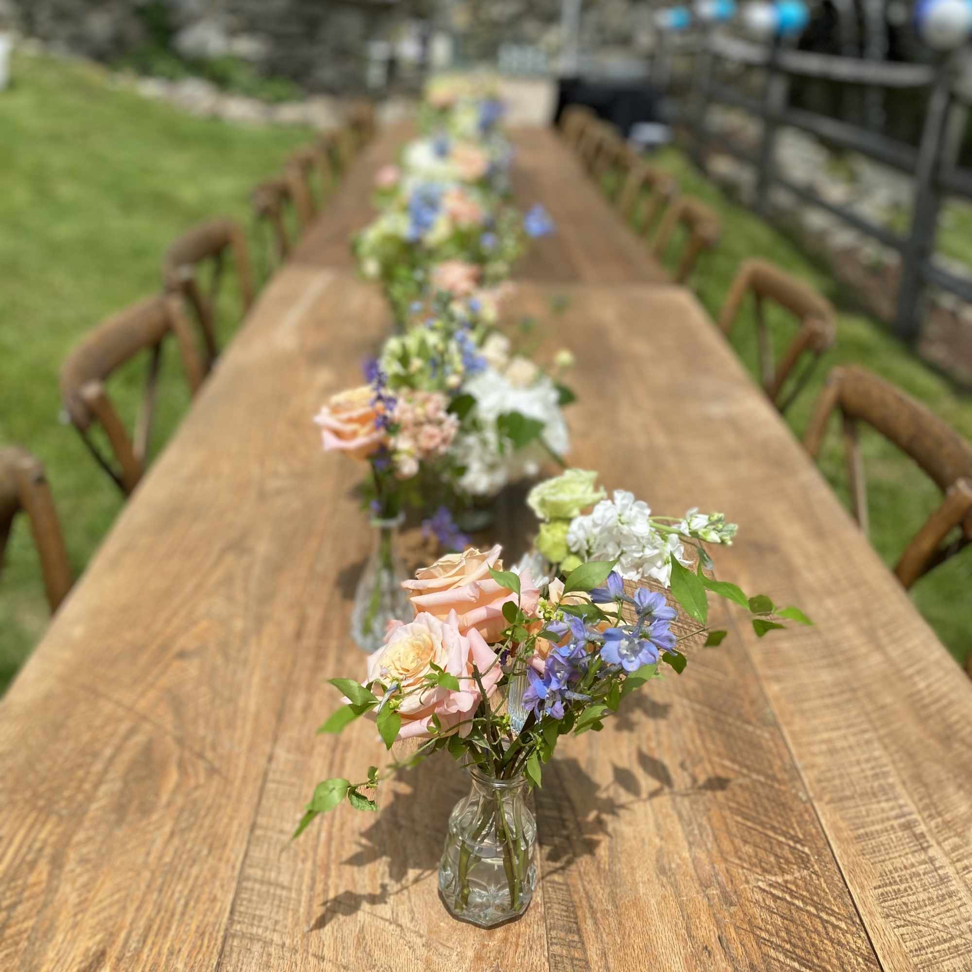 Small floral arrangements in clear glass vases line a wooden table outdoors.