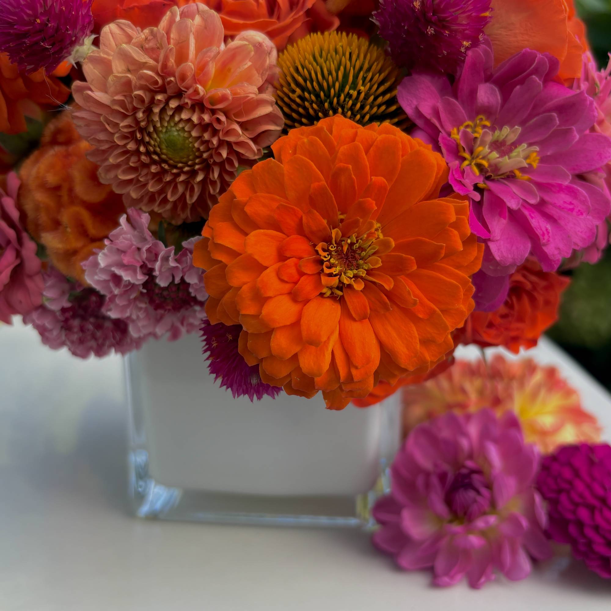 Bright mixed bouquet of orange, pink, and purple flowers in a clear square vase