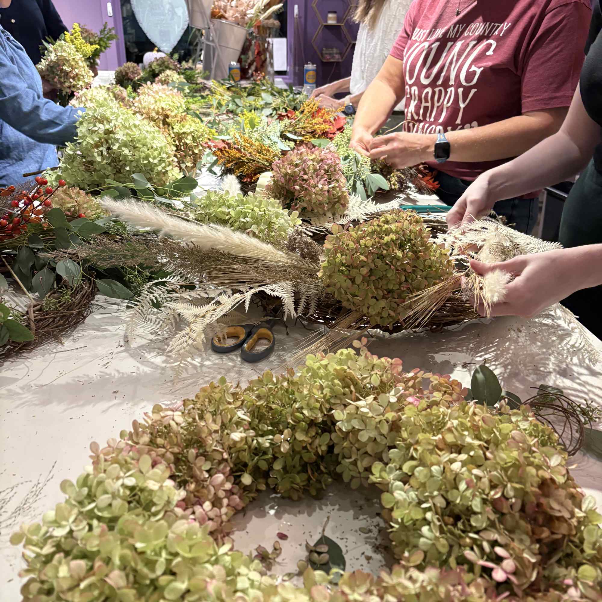 People arranging floral wreaths and hydrangea pieces on a table in a shop