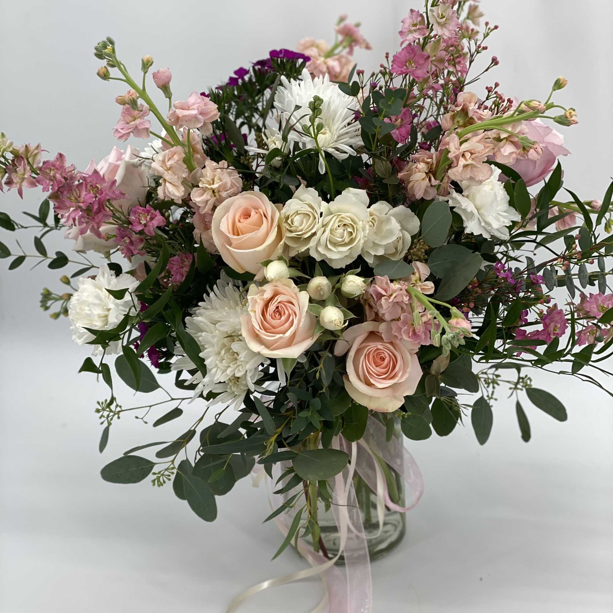 Tall arrangement of peach roses, white mums, and pink blooms in a glass vase with trailing ribbons