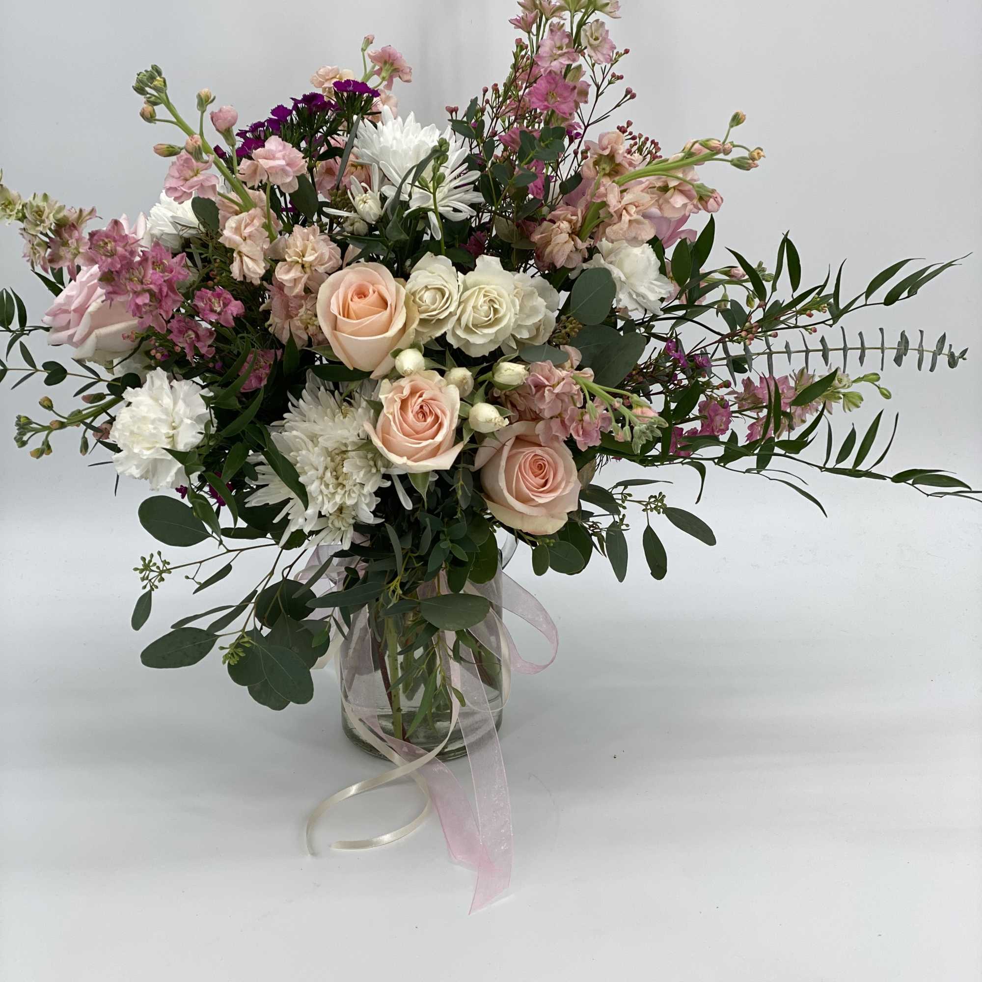 Mixed arrangement of peach roses, white blooms, and pink flowers in a clear glass vase with ribbon