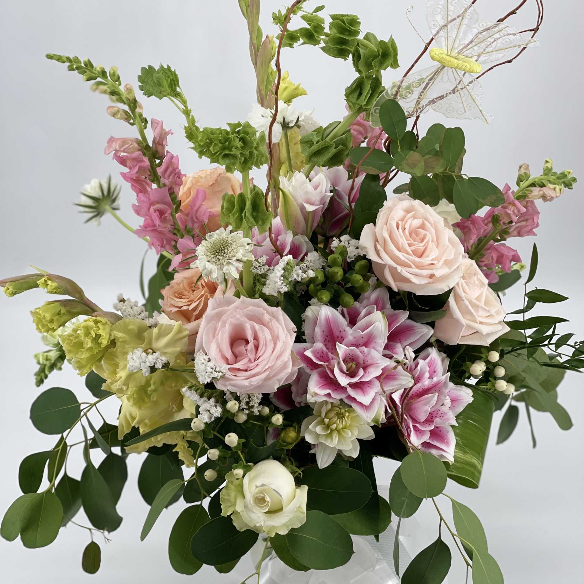 Tall pastel arrangement with pink roses, lilies, green bells of Ireland and a butterfly pick in a glass vase.