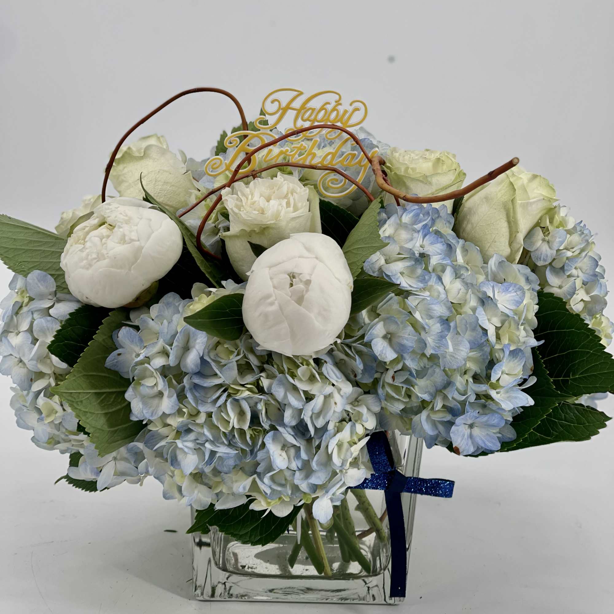 Blue hydrangea and white peony arrangement in a glass vase with a birthday topper