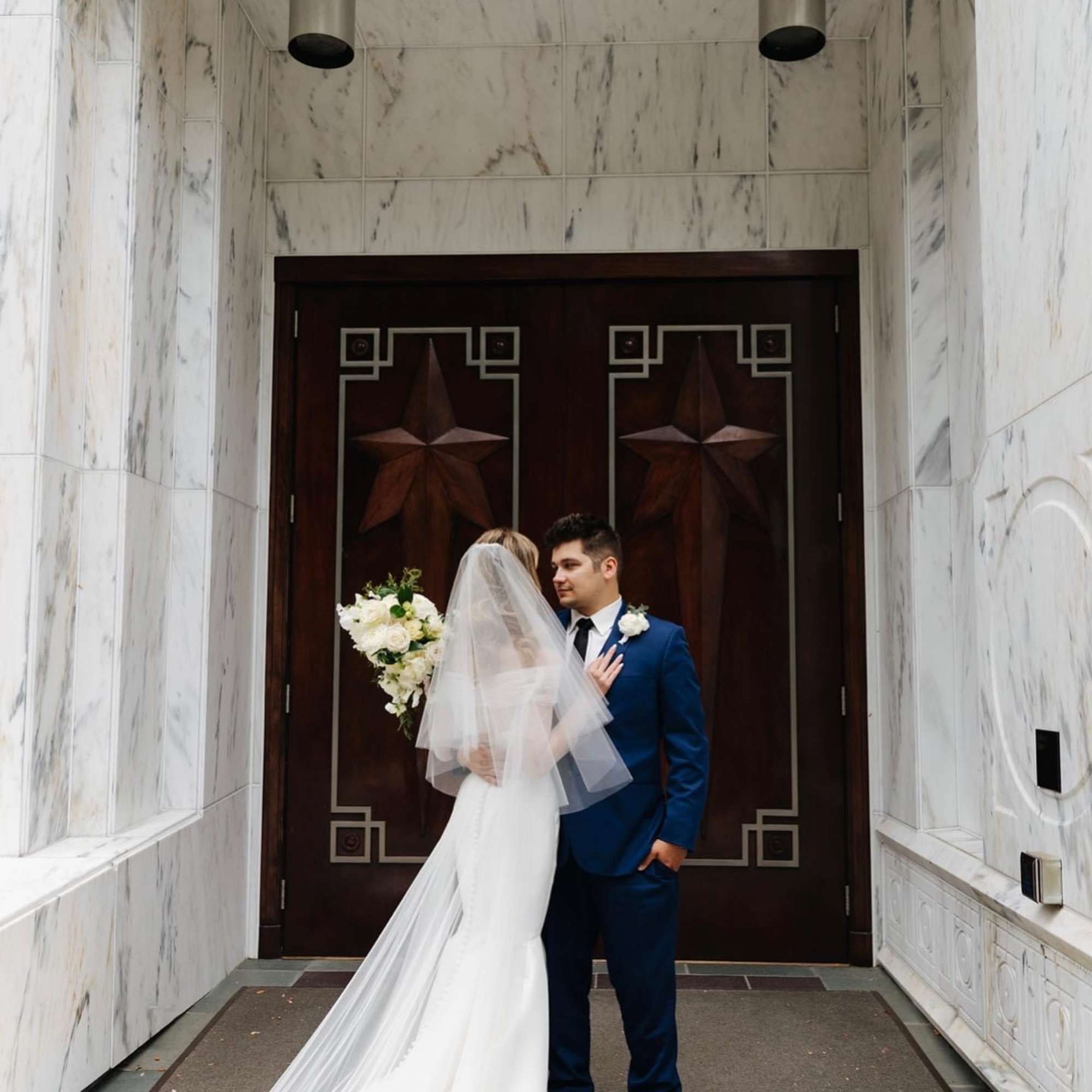 Bride and groom posing with a white bouquet in front of dark wooden doors