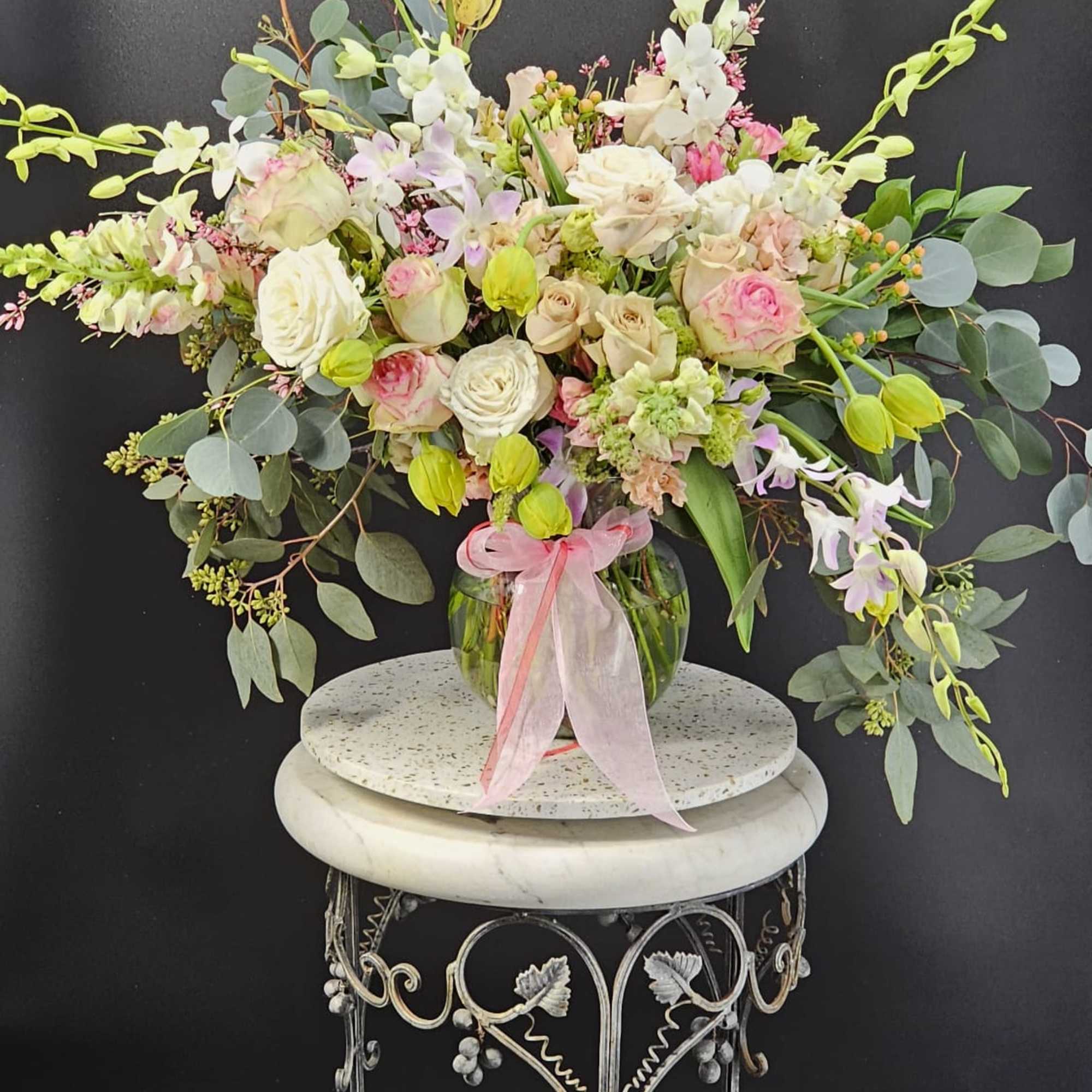 Large pastel arrangement of roses and orchids in a glass vase with ribbon on a decorative pedestal stand