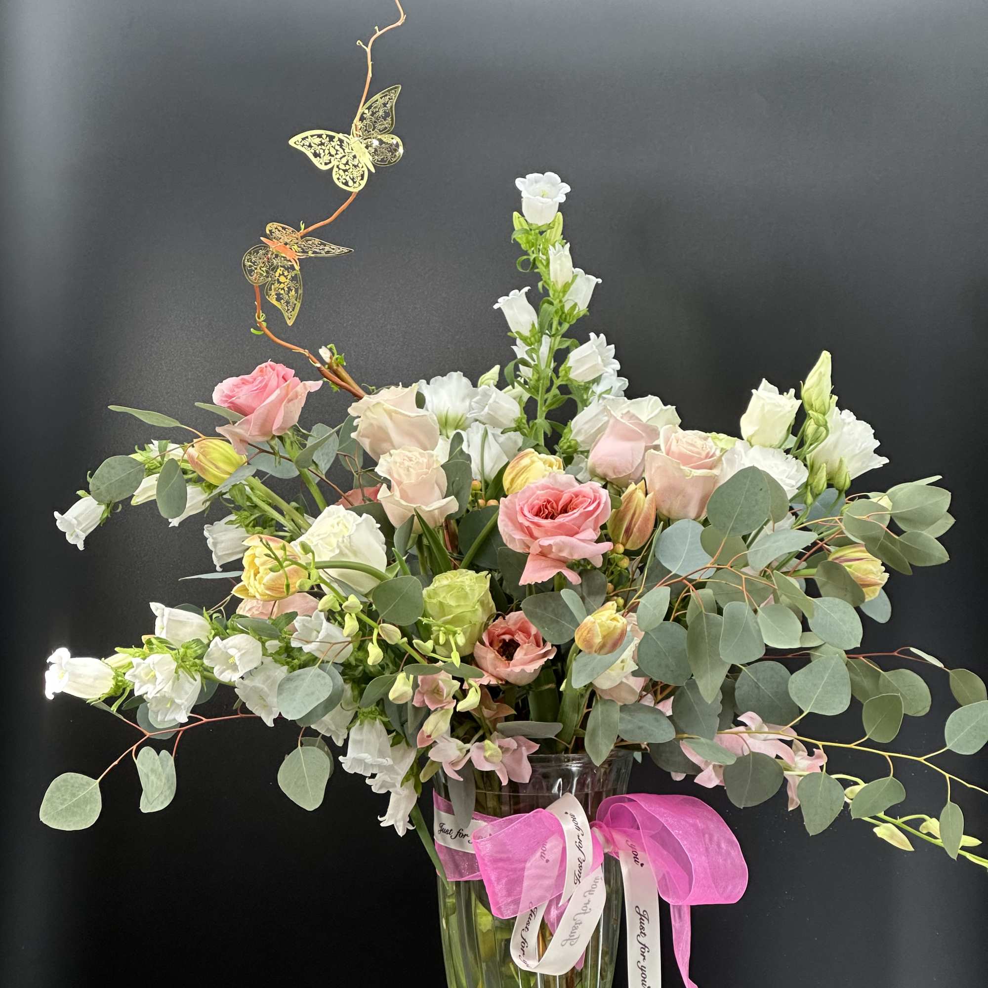 Tall pastel arrangement of pink and white roses and lisianthus in a clear glass vase with butterfly accents.