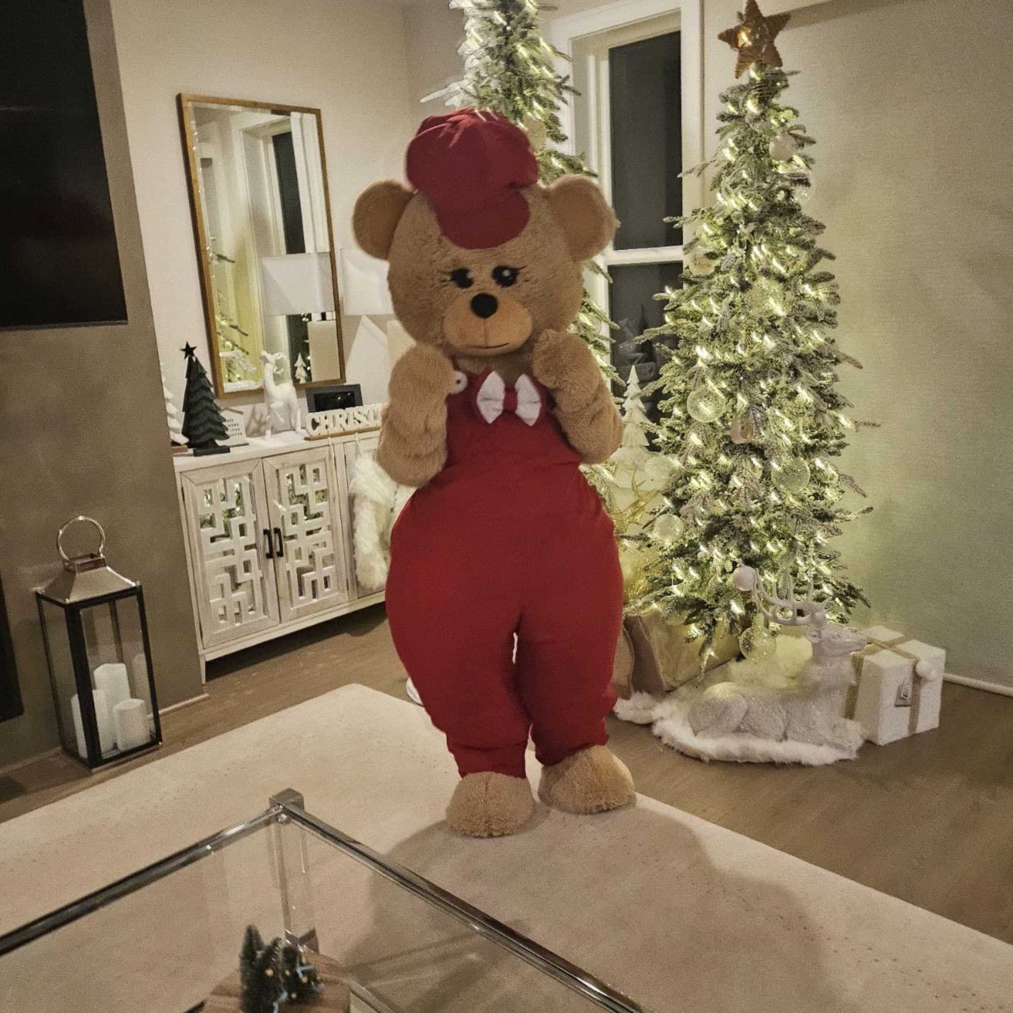 Life-size teddy bear in red overalls and cap standing in a Christmas-decorated living room