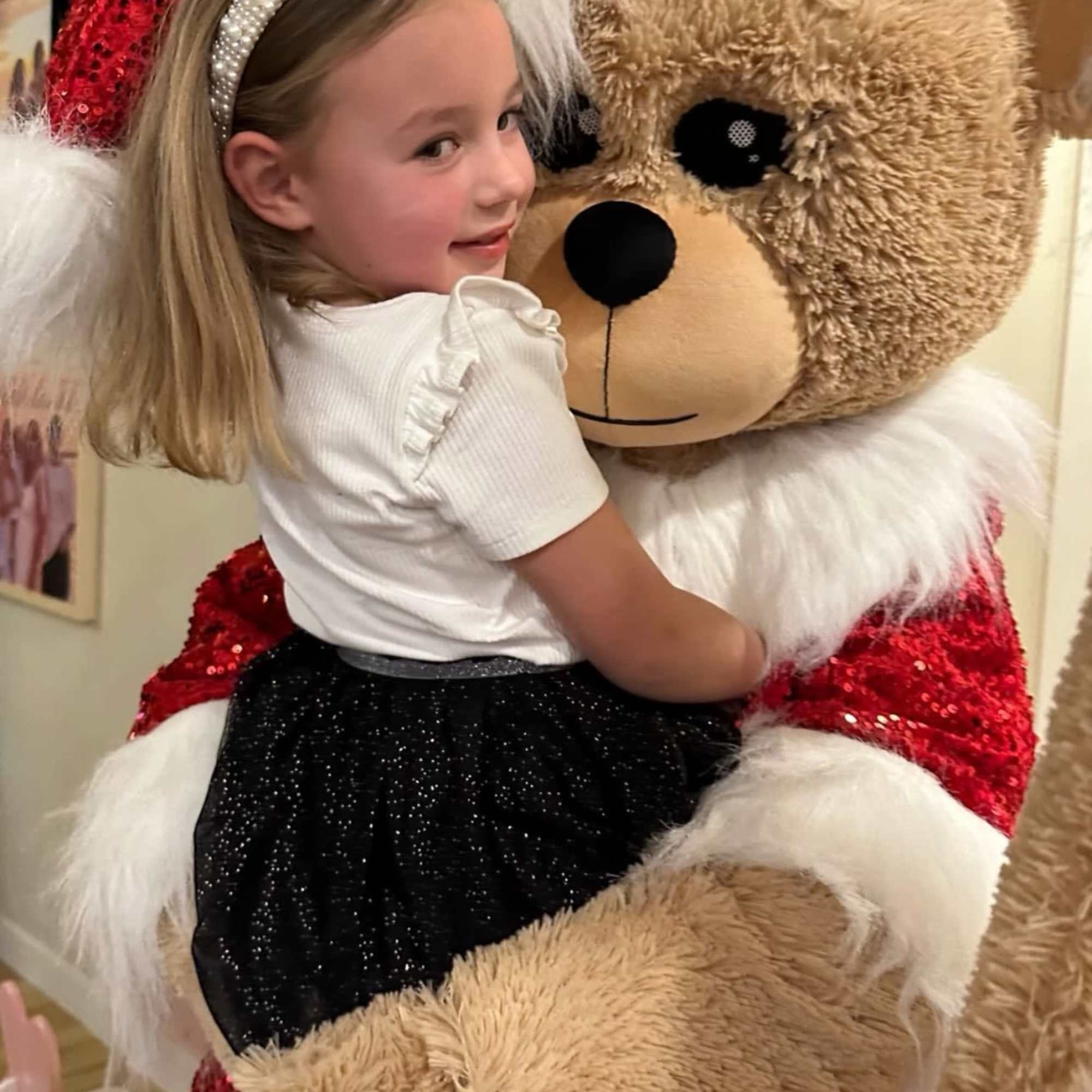 Young child hugging an oversized teddy bear dressed in a red sequin Santa outfit.