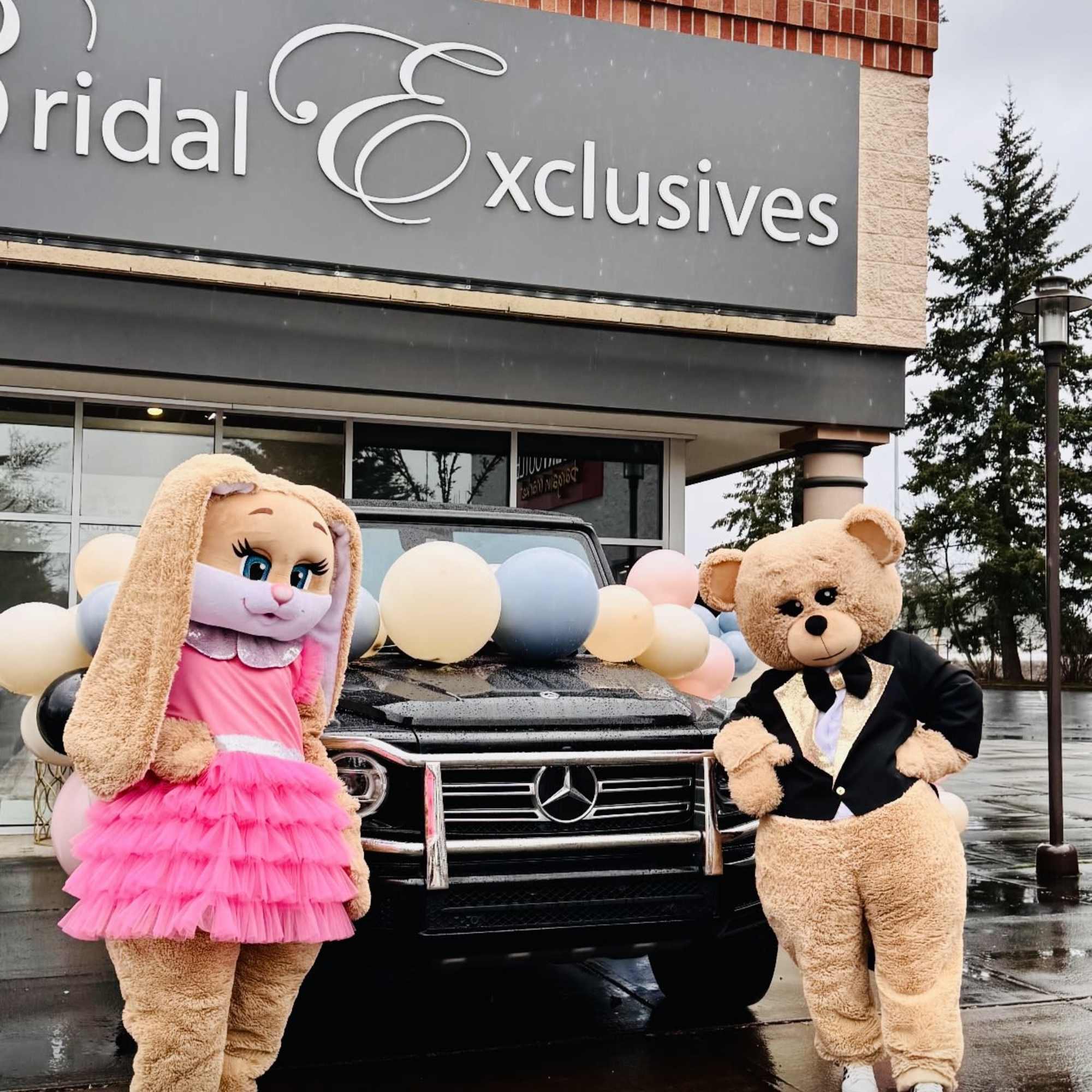 Costumed bunny and bear stand by a black SUV decorated with pastel balloons outside a bridal shop.