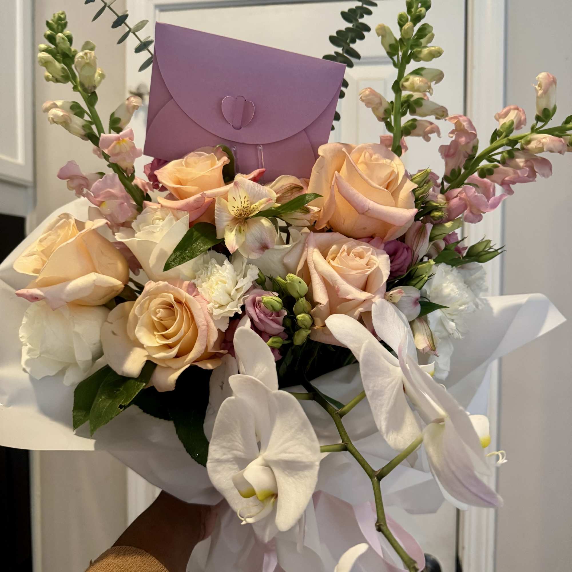 Handheld bouquet of peach roses, white orchids, and pink snapdragons wrapped in white paper