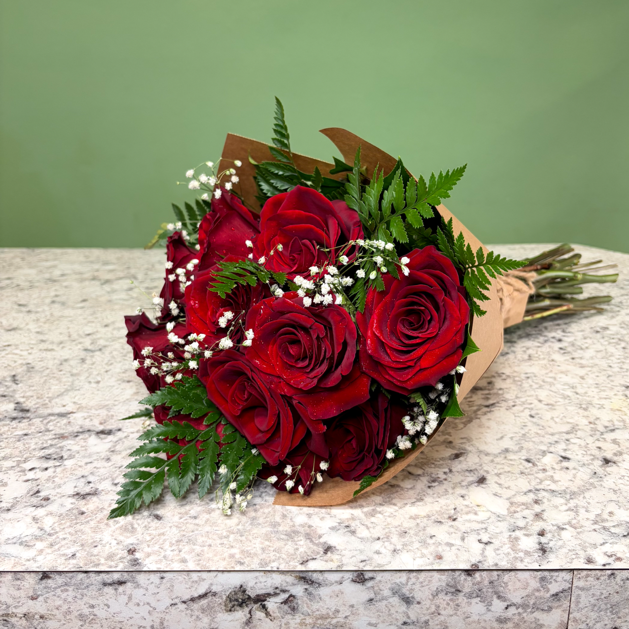 Hand-tied bouquet of deep red roses with white filler flowers wrapped in brown paper on a stone counter