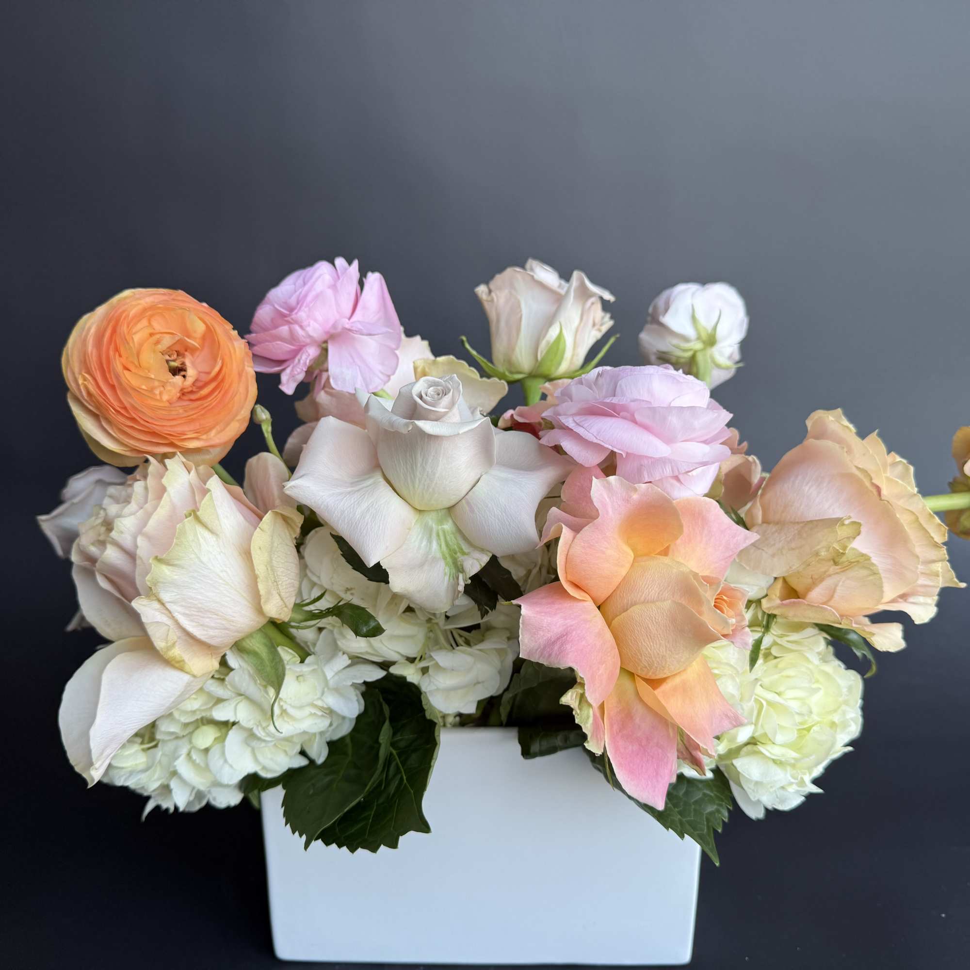 Low arrangement of peach, pink, and white roses and ranunculus in a white rectangular vase