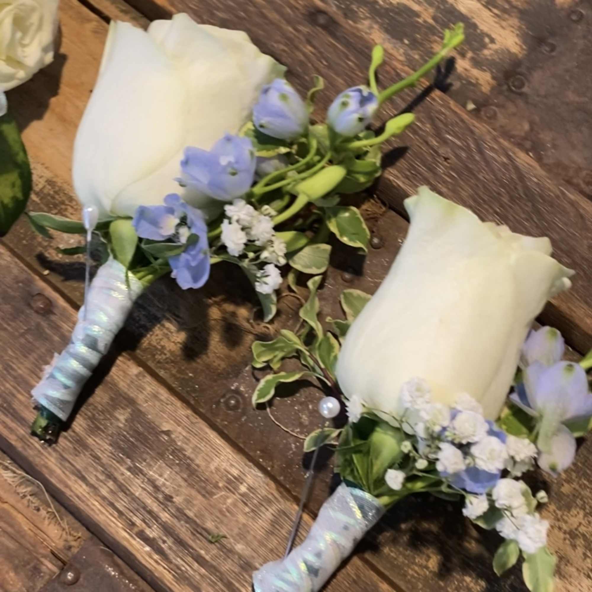 Two small white rose boutonnieres with blue flowers on a wooden surface