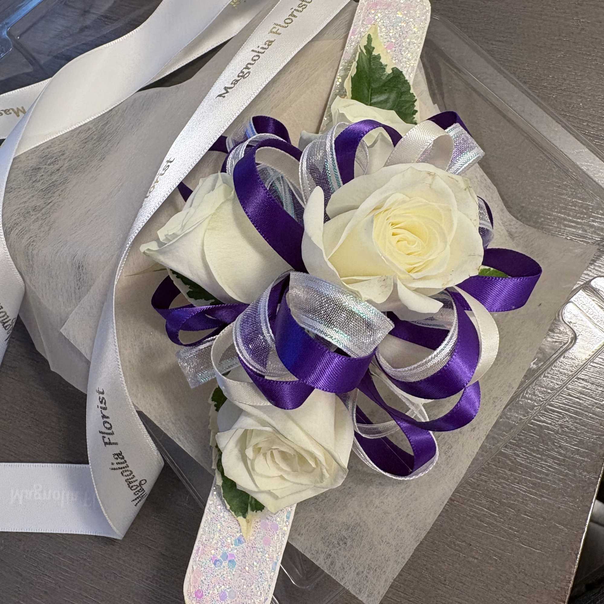 Three white roses with purple ribbon accents in clear packaging