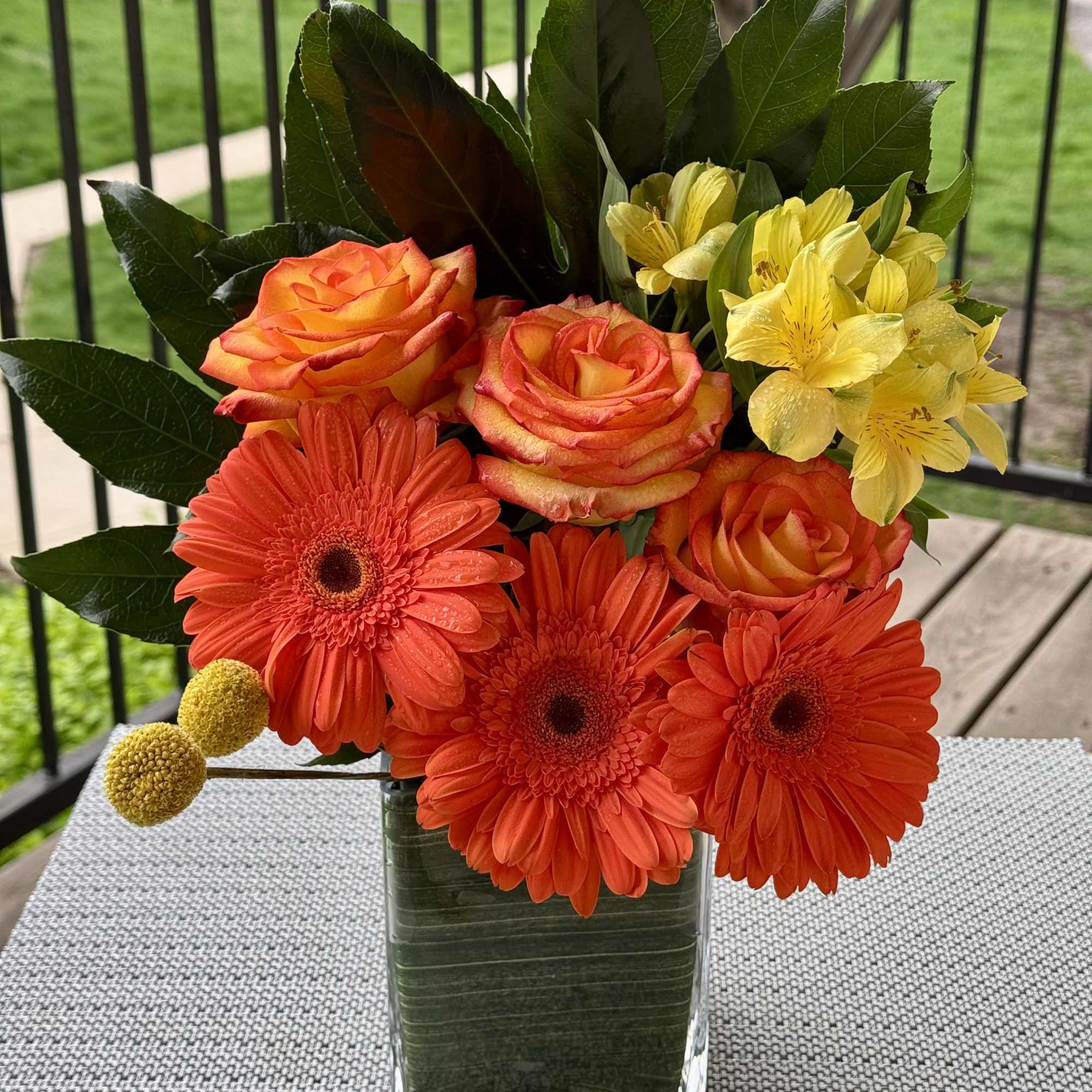 Orange roses and gerbera daisies in a clear glass vase with yellow flowers