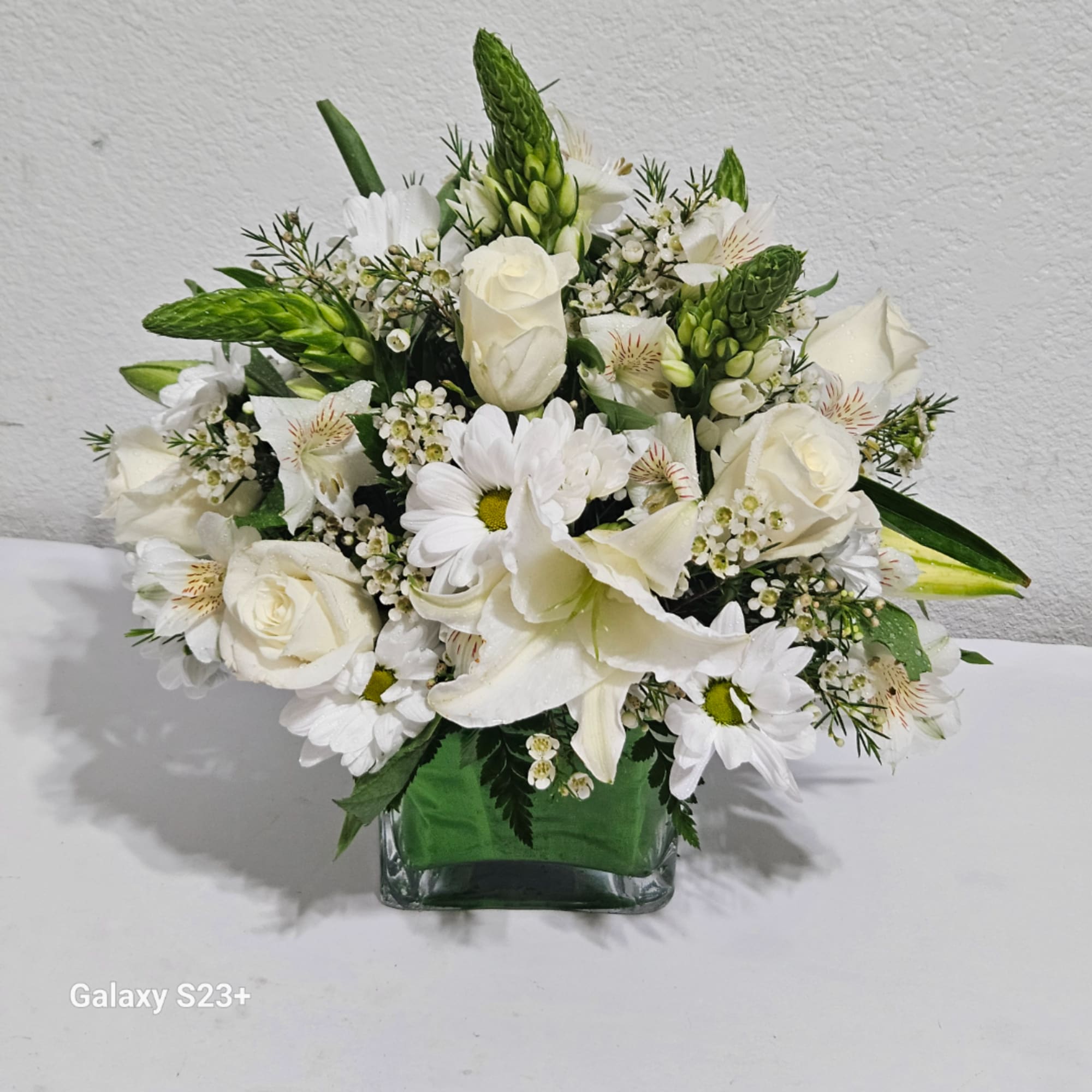 White floral arrangement with roses, lilies, and daisies in a square glass vase