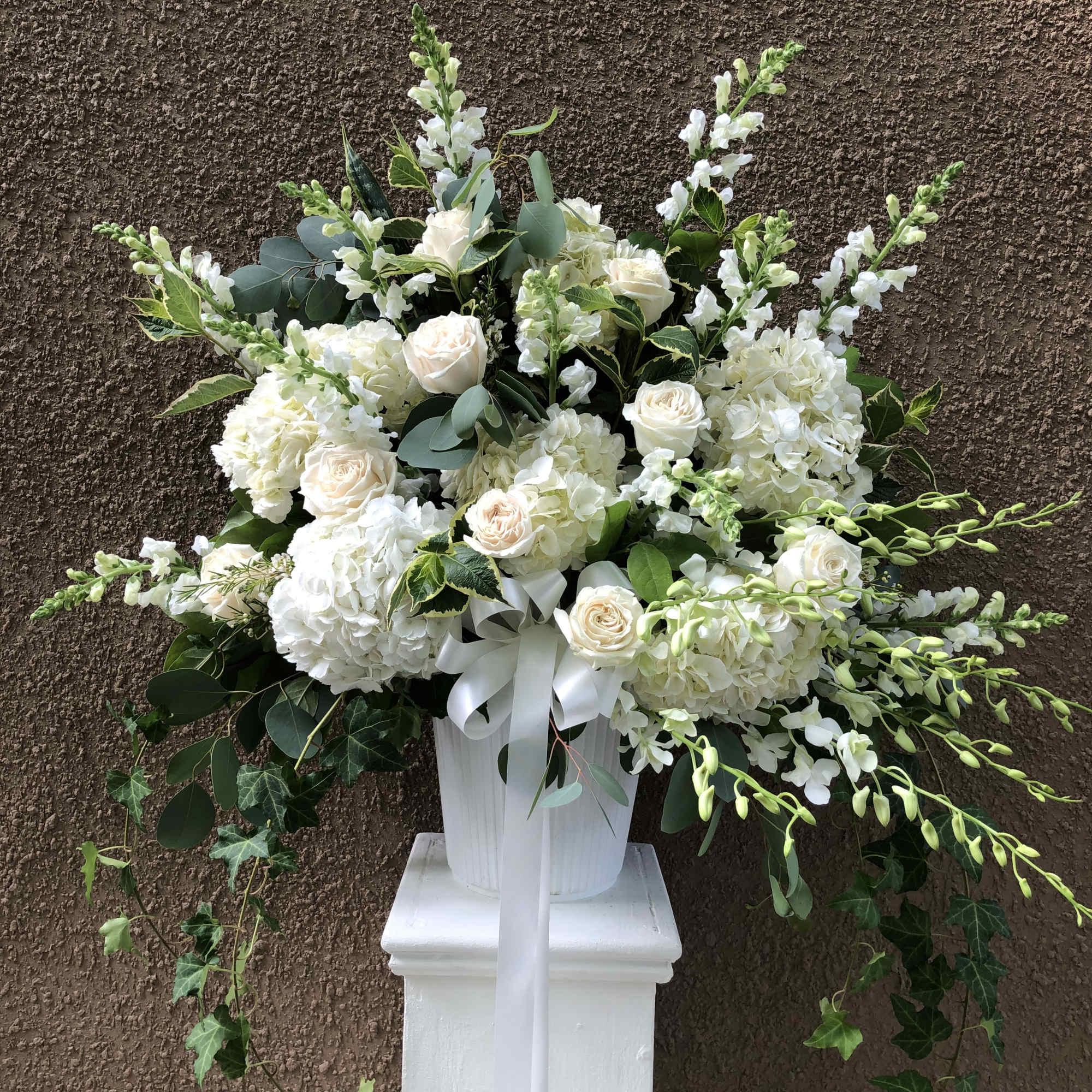 White floral arrangement in a white pedestal vase with a ribbon bow