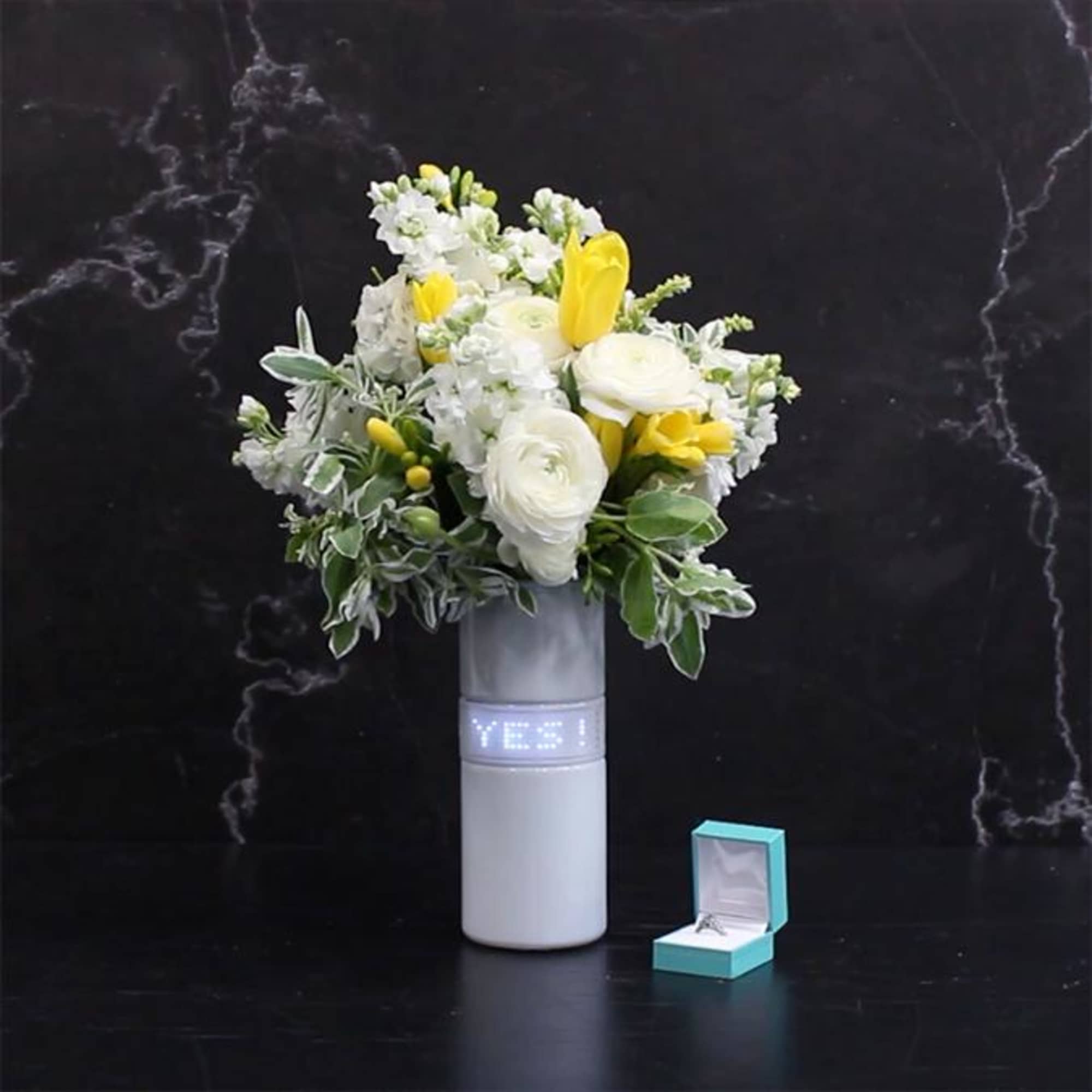 White and yellow floral arrangement in a tall vase with a small ring box beside it