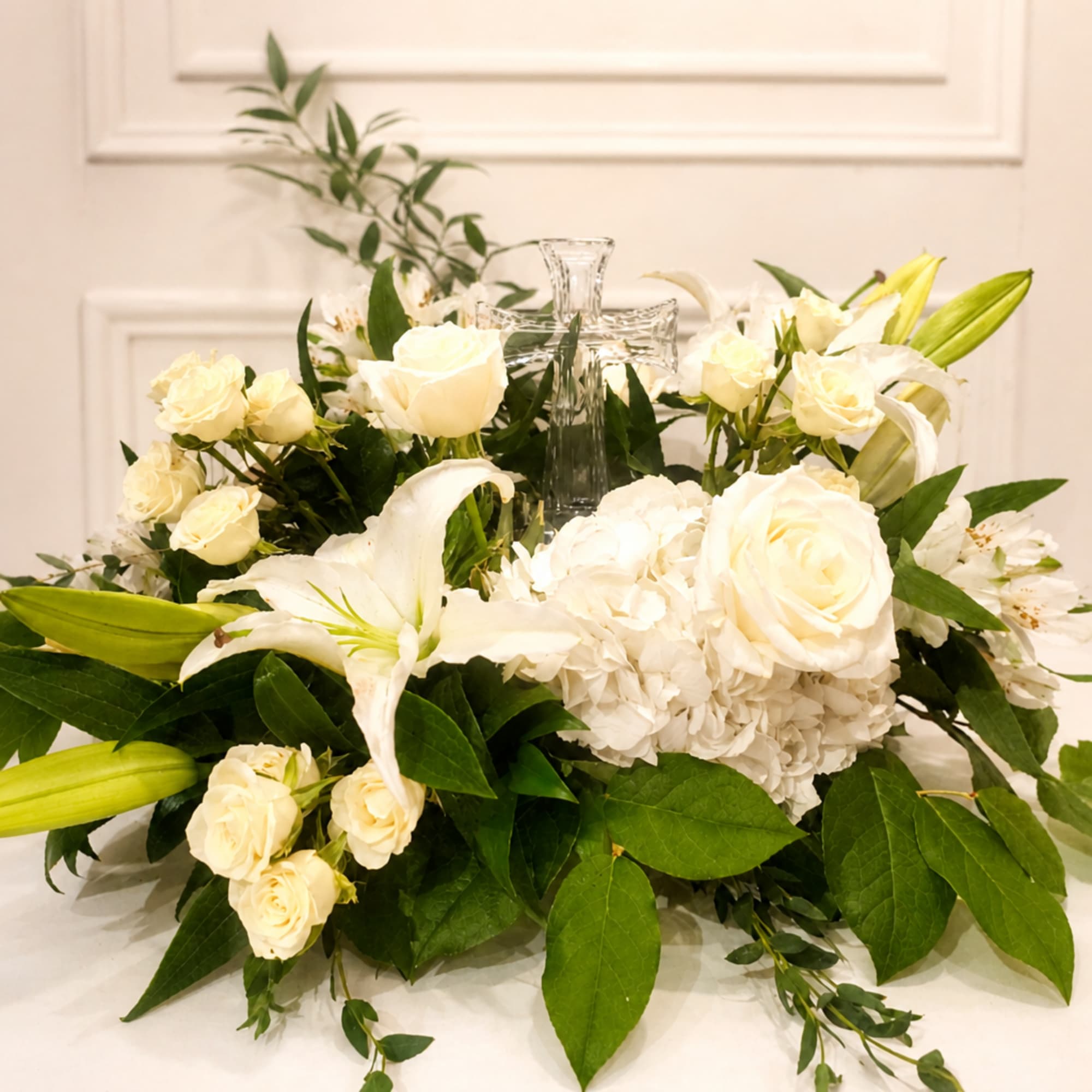 White floral arrangement with roses, lilies, and a clear glass cross centerpiece