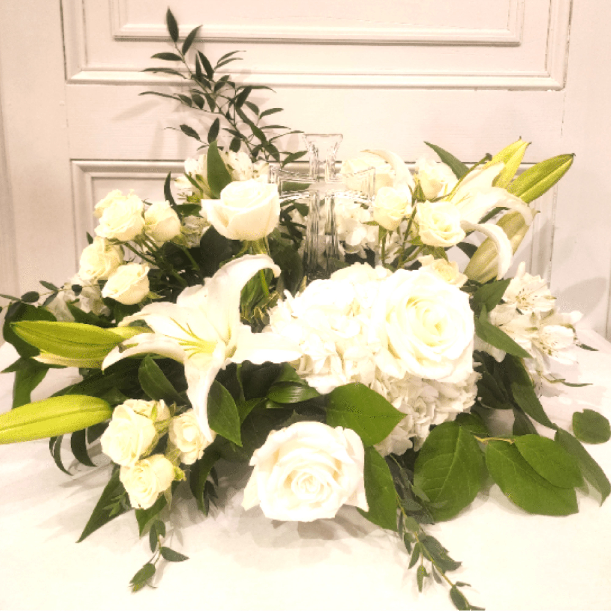 White lilies, roses, and hydrangea surround a keepsake glass cross. Classic and