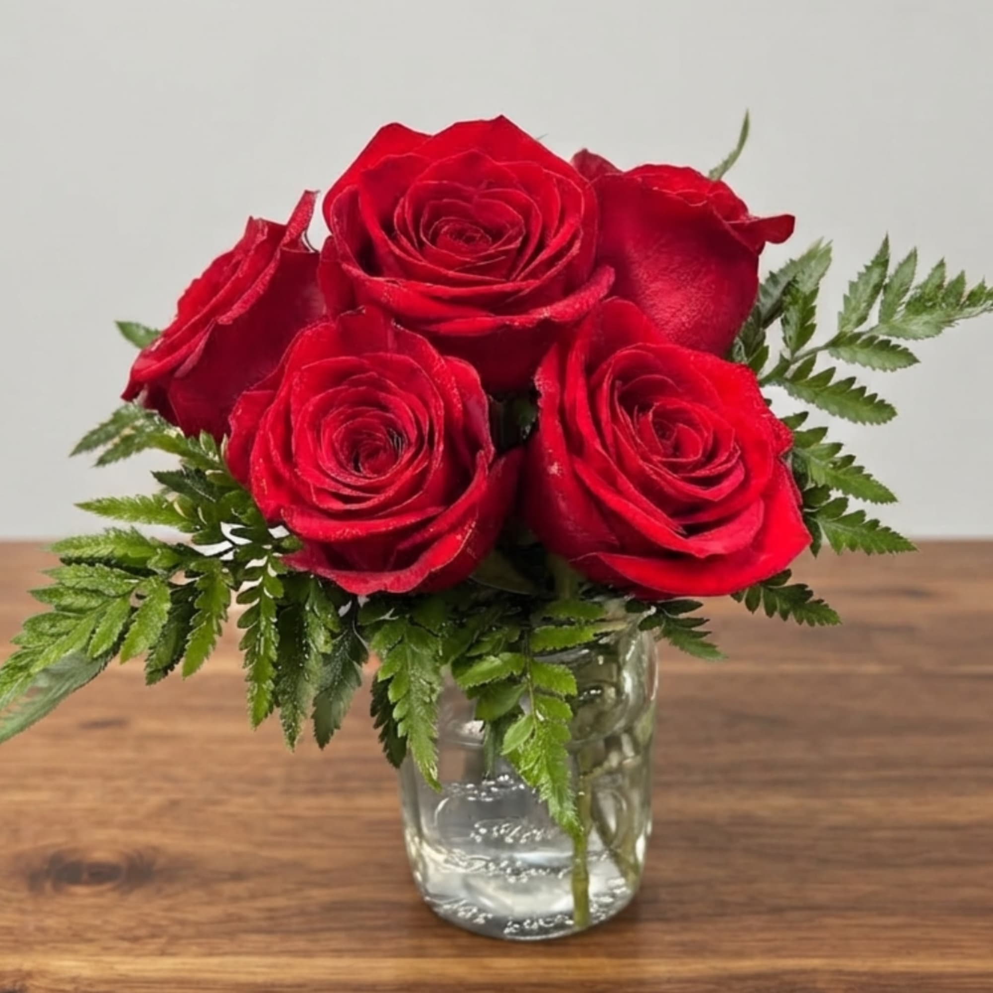 Red roses arranged in a clear glass jar with fern foliage