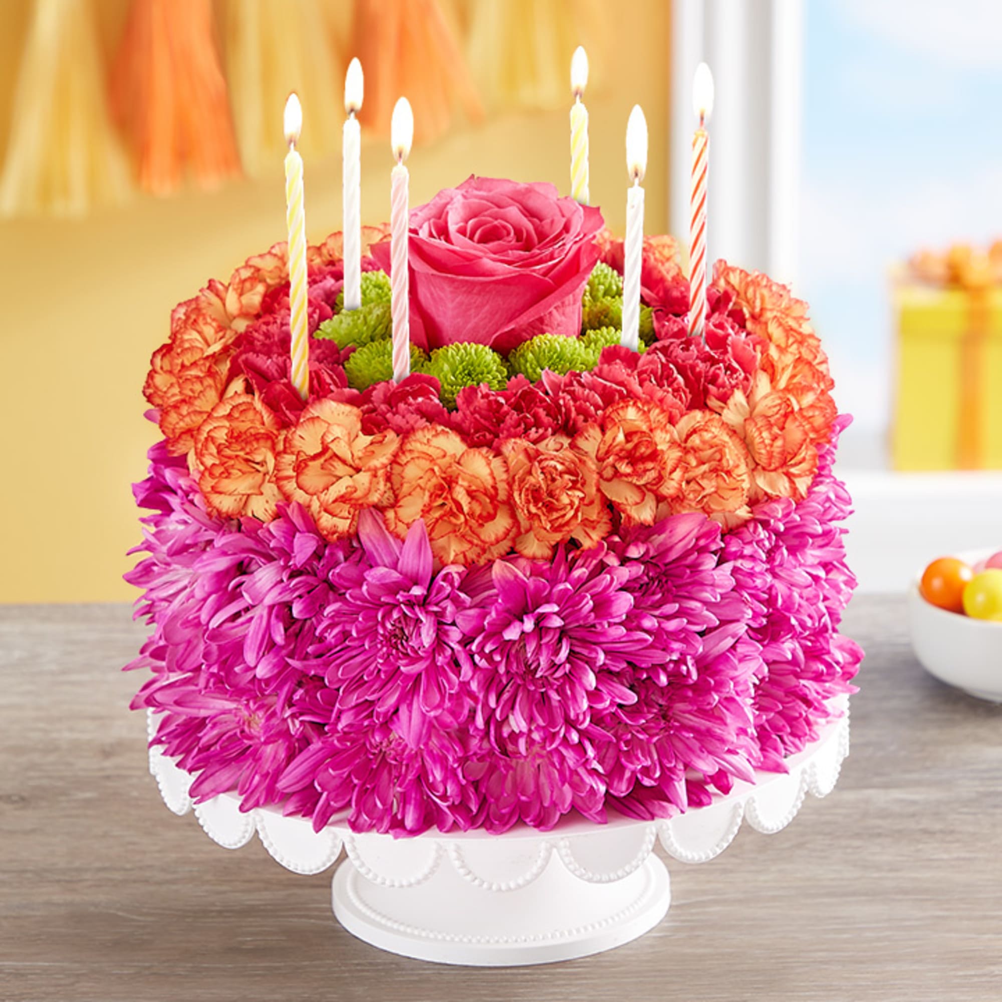 Round floral cake of pink and orange blooms with a central rose and lit birthday candles on a white stand
