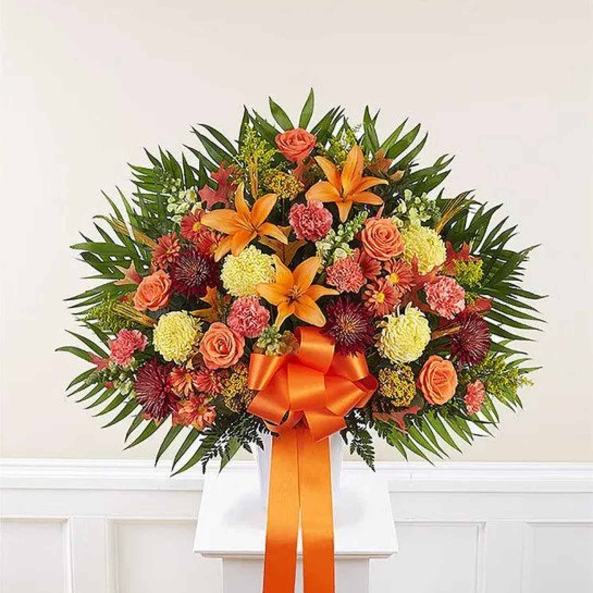 Fan-shaped arrangement of orange lilies, roses, carnations and mums with an orange bow in a white container