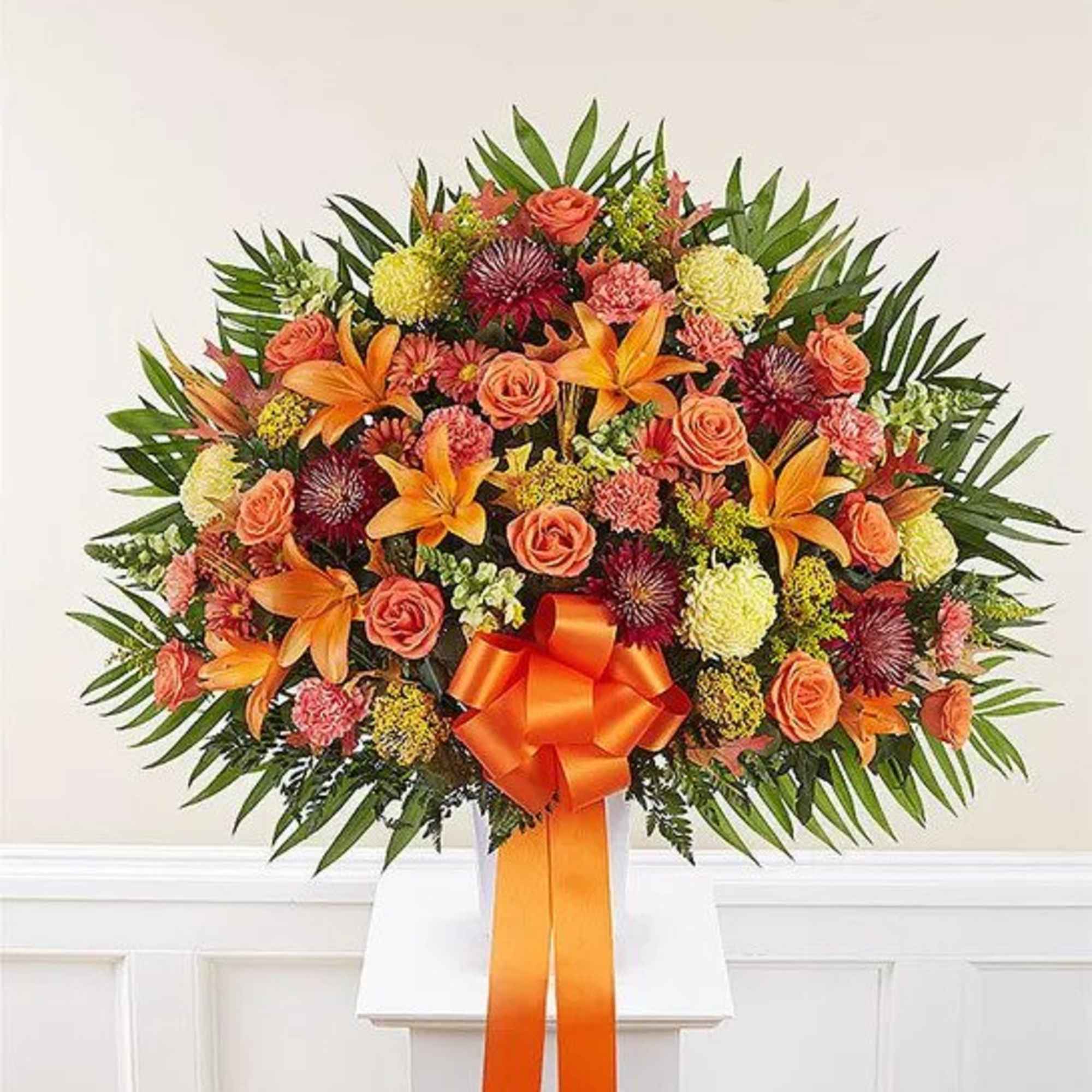 Large orange and yellow floral arrangement with roses and lilies in a white container with an orange bow
