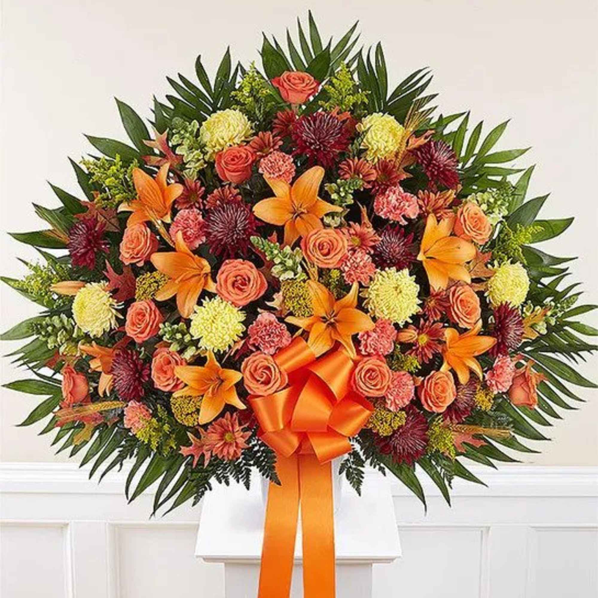 Large standing spray of orange lilies, peach roses, and yellow and burgundy mums with an orange bow