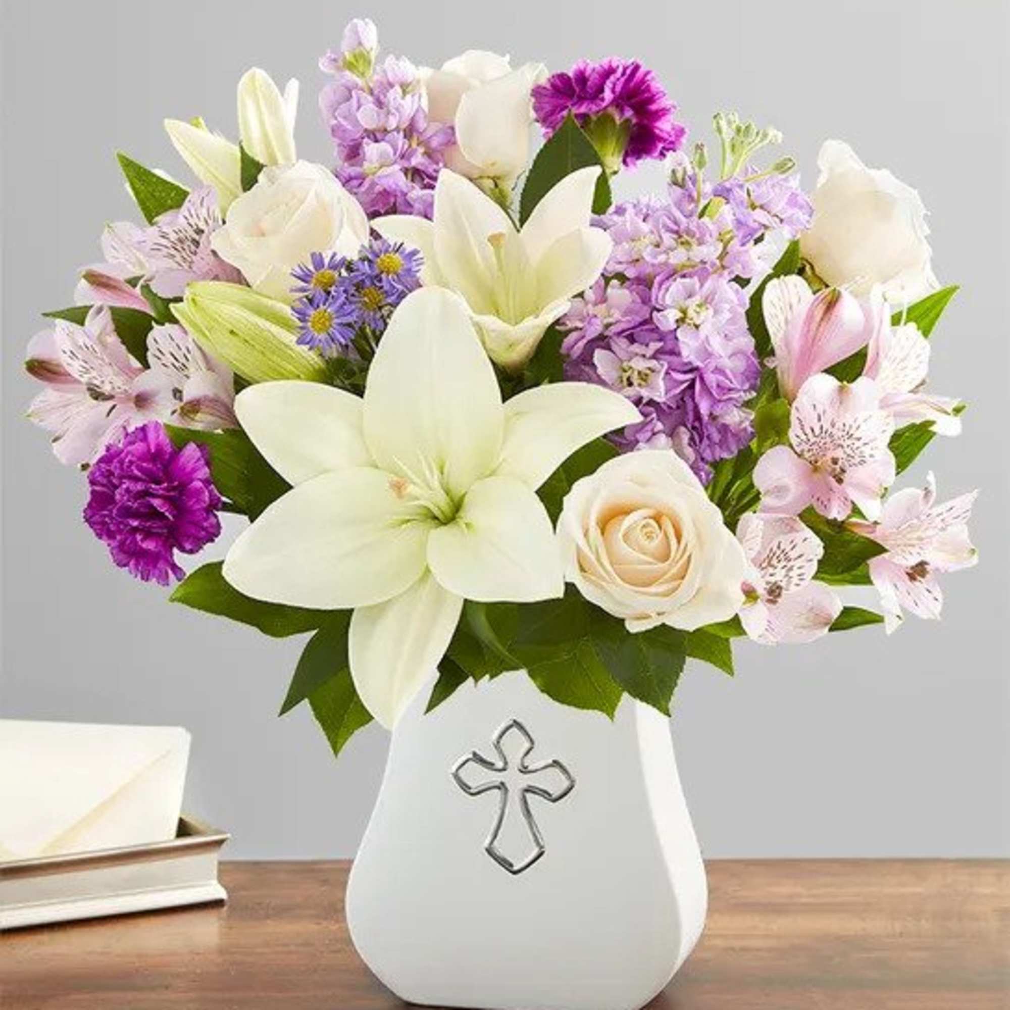 White lilies, roses and purple blooms in a white vase with a silver cross on a wooden table