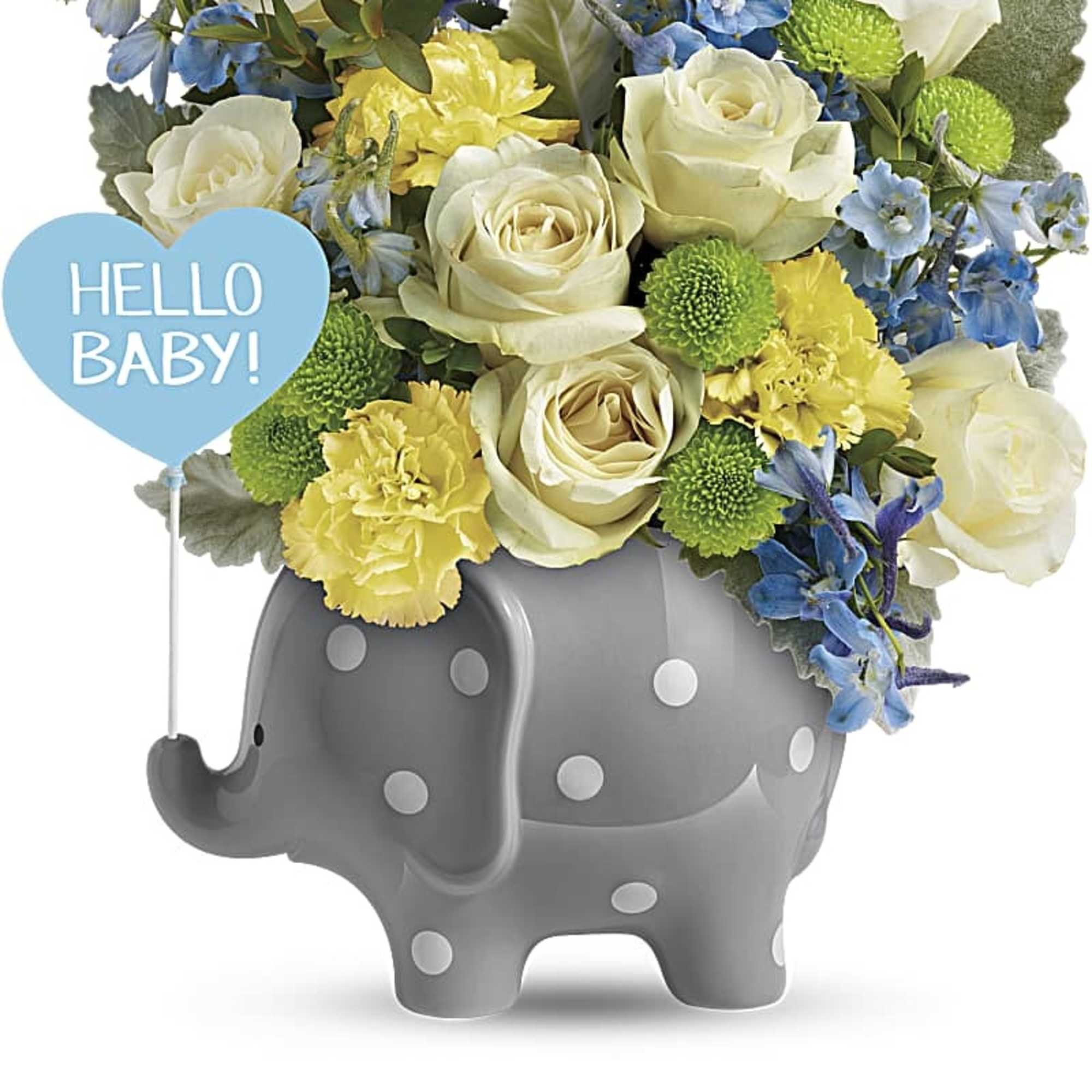 Cream roses, yellow carnations, and blue flowers in a gray polka-dot elephant vase with a Hello Baby pick.