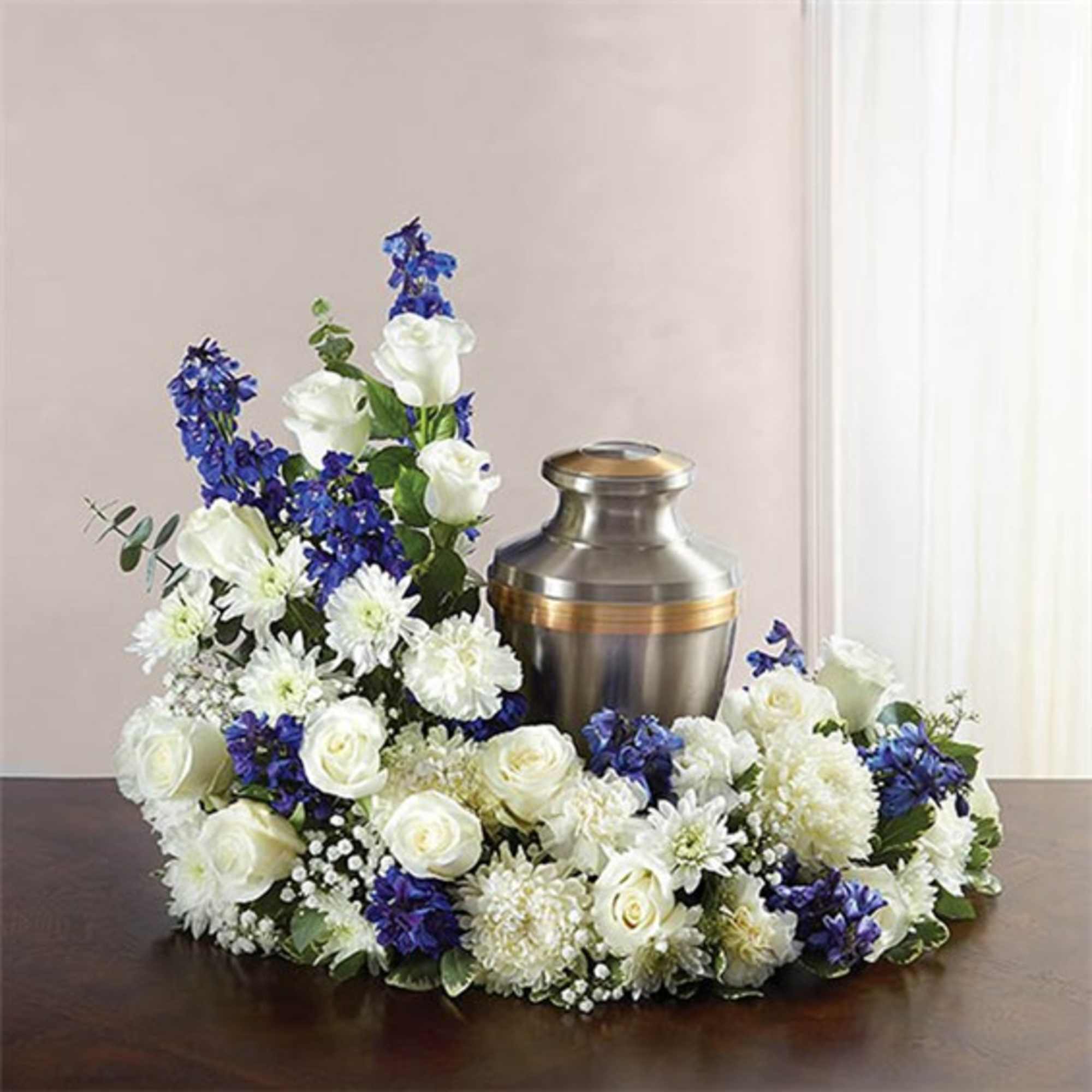 White roses and mums with blue delphinium arranged in a ring around a metal cremation urn